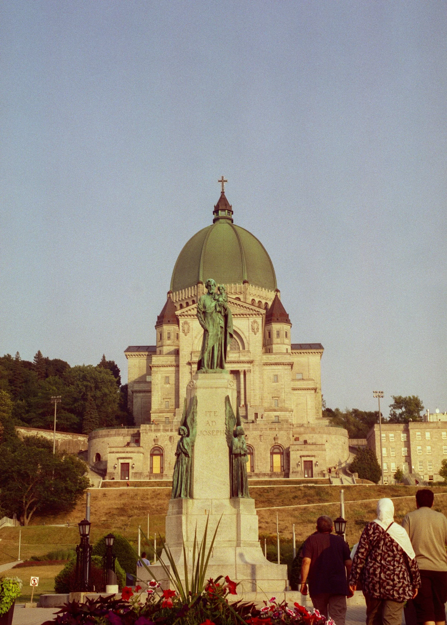 View of a domed church building with a statue of a religious figure in the foreground, people walking nearby, and flowers in the foreground.