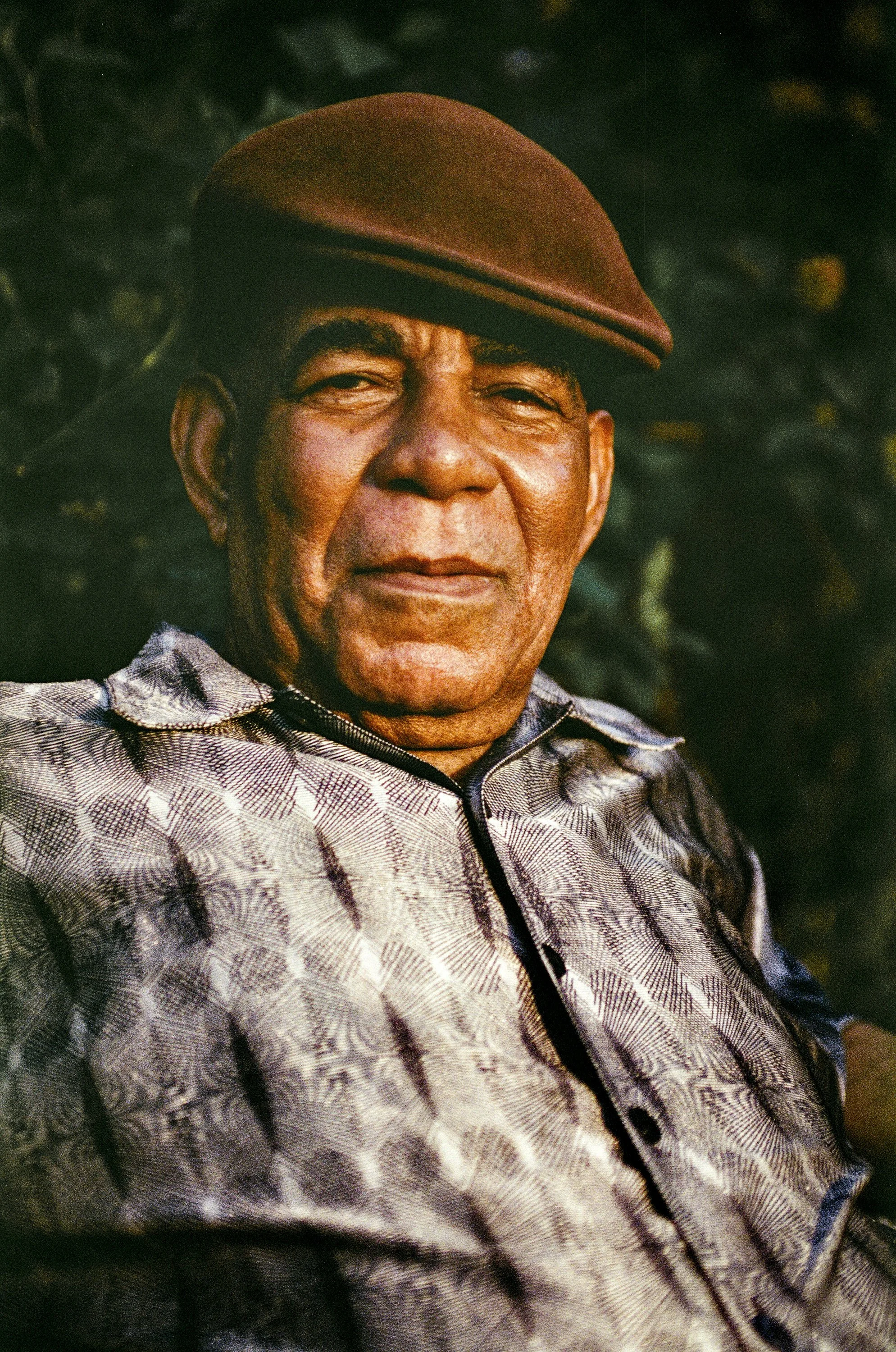 A close-up portrait of an elderly man wearing a brown beret and a patterned shirt, outdoors against a blurred background of dark foliage.