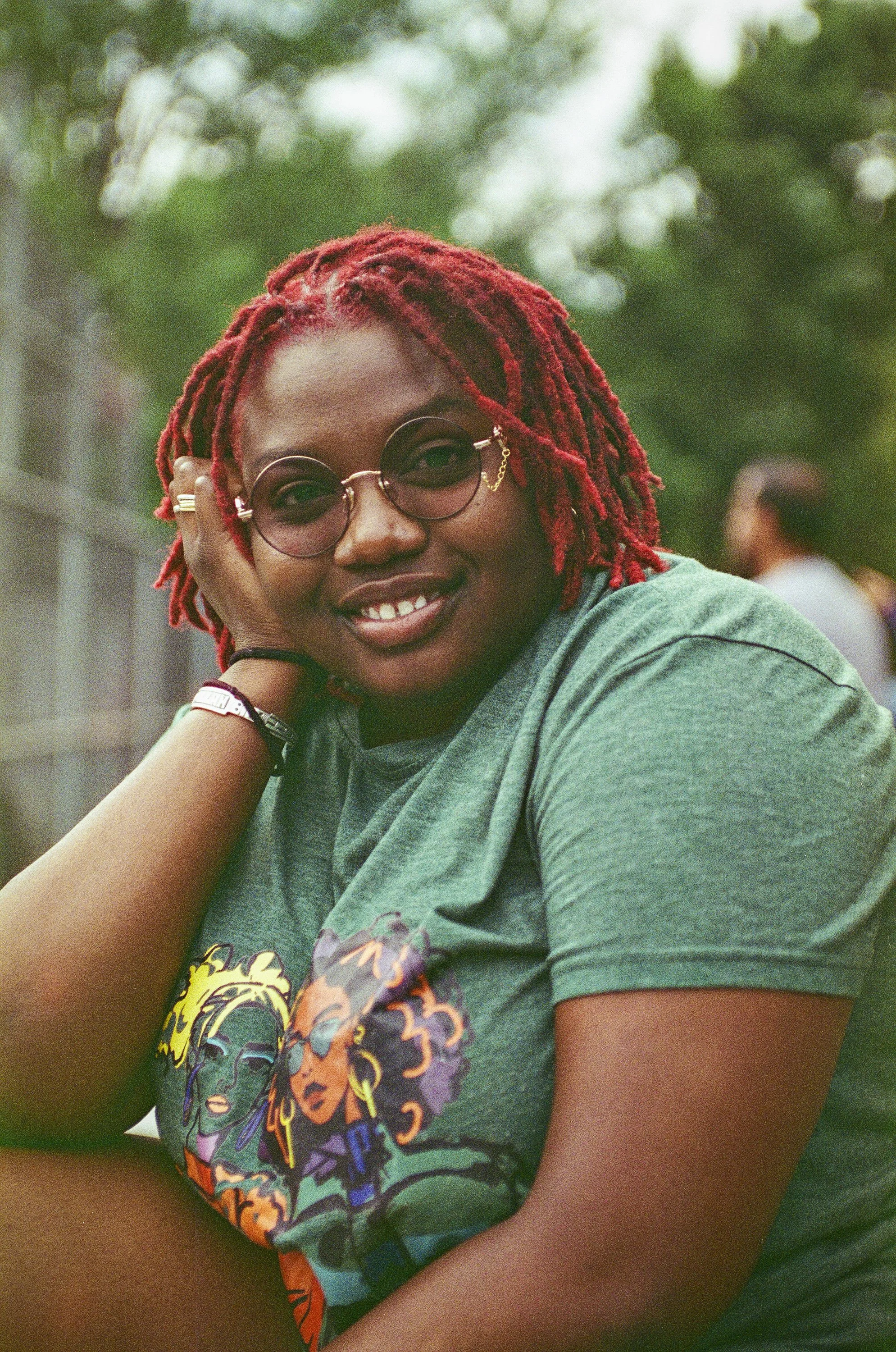 A woman with red dreadlocks wearing sunglasses and a gray T-shirt with a colorful graphic design, sitting outdoors with green blurry background.