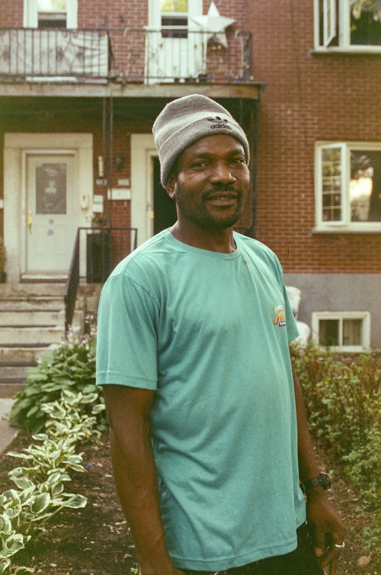 A man with dark skin wearing a gray beanie and a teal T-shirt standing outside in front of a red brick building with a small garden bed in the foreground.