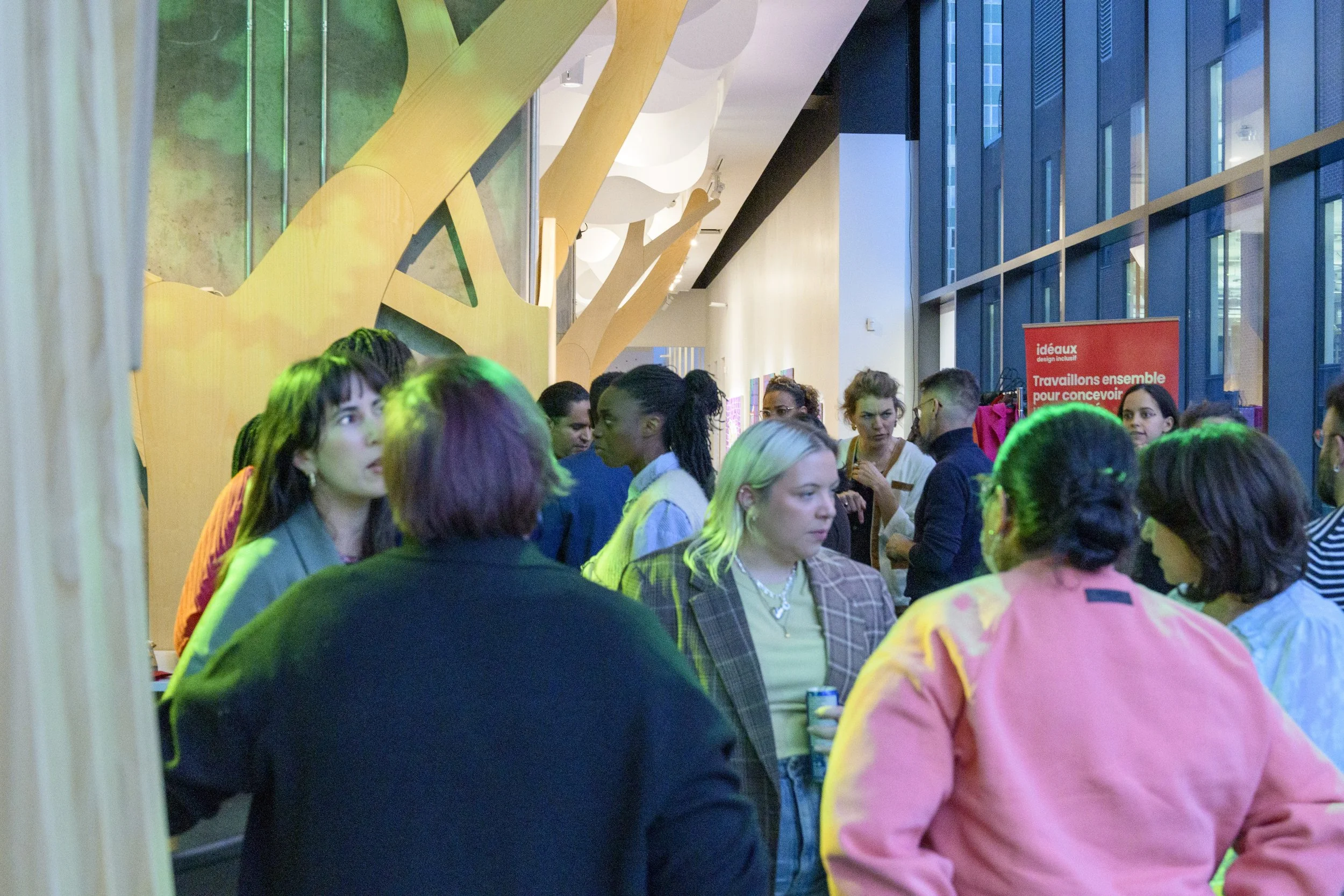 A group of diverse people gathered indoors, some engaging in conversation and others looking around, with large windows on the right side and colorful lighting.