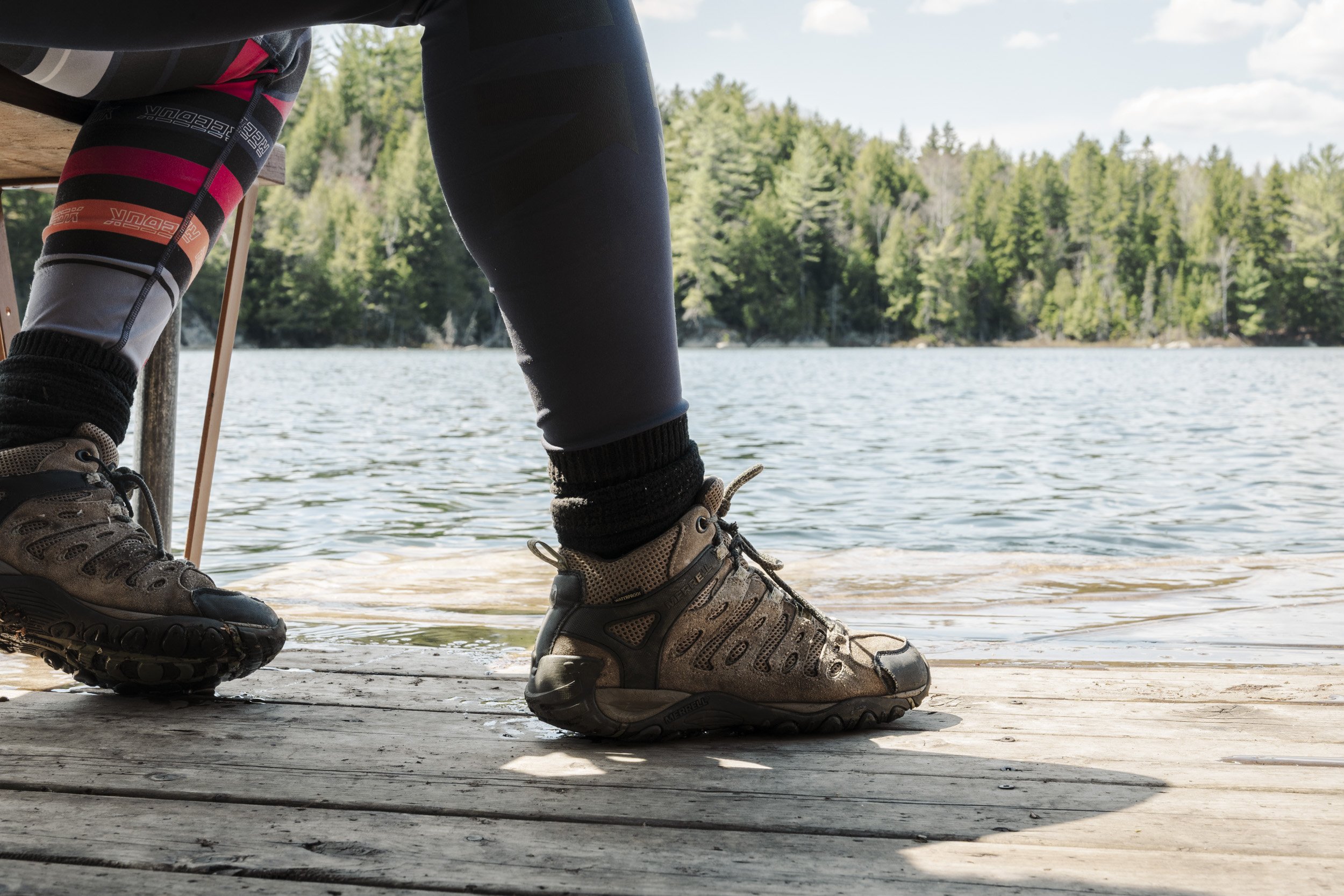 Close-up of hiking boots and lower legs of a person sitting on a dock by a lake, with a forest in the background. The person is wearing outdoor gear, including patterned leggings and a jacket.