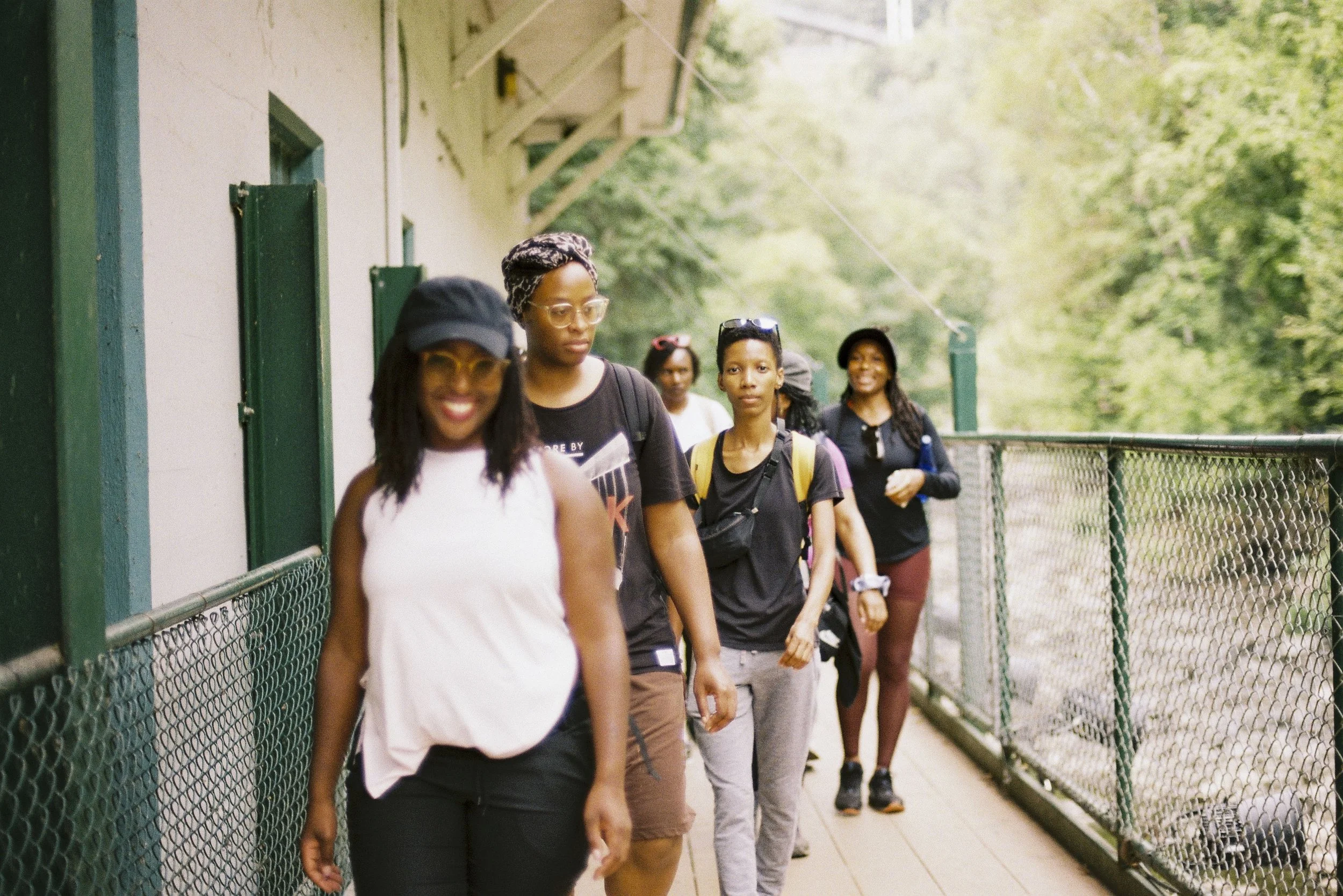 A group of diverse women walking along a fenced outdoor pathway surrounded by greenery on a sunny day.