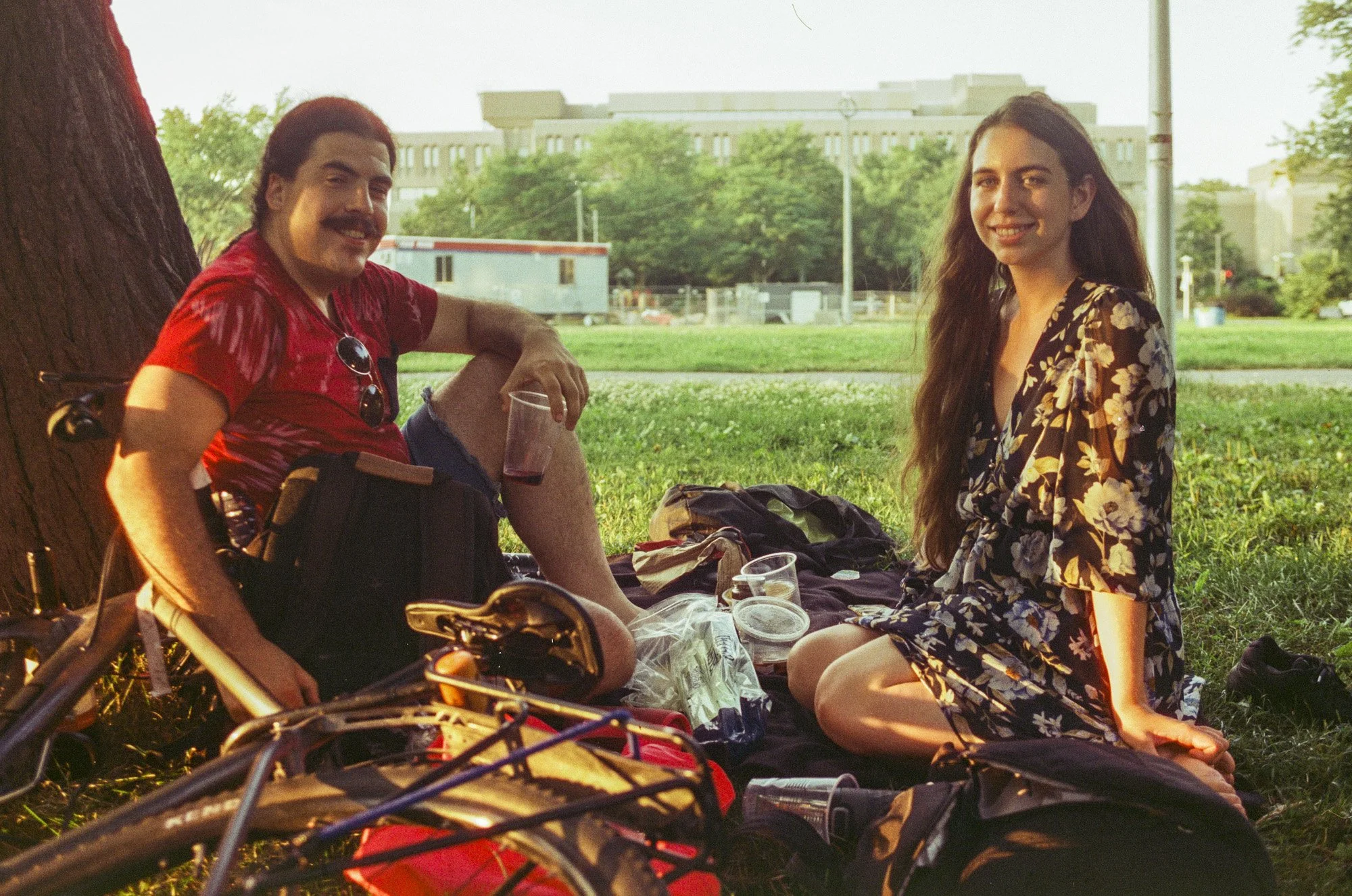A man and woman sitting outdoors on grass, smiling at the camera, with trees and buildings in the background during sunset.