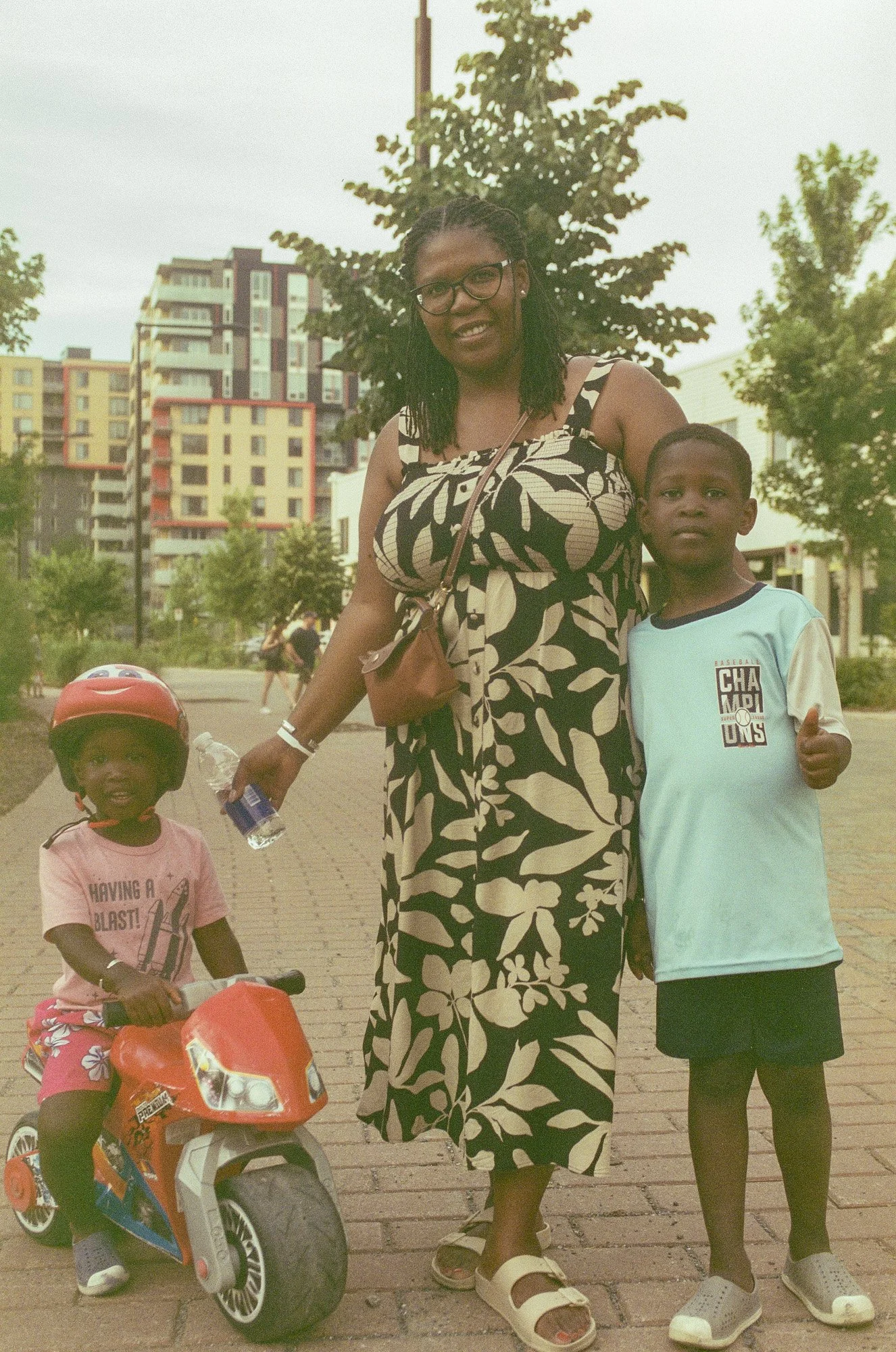 A woman with glasses, dreadlocks, and a floral dress is standing outdoors with two children, one on a toy motorcycle, in an urban park setting with trees and apartment buildings in the background.
