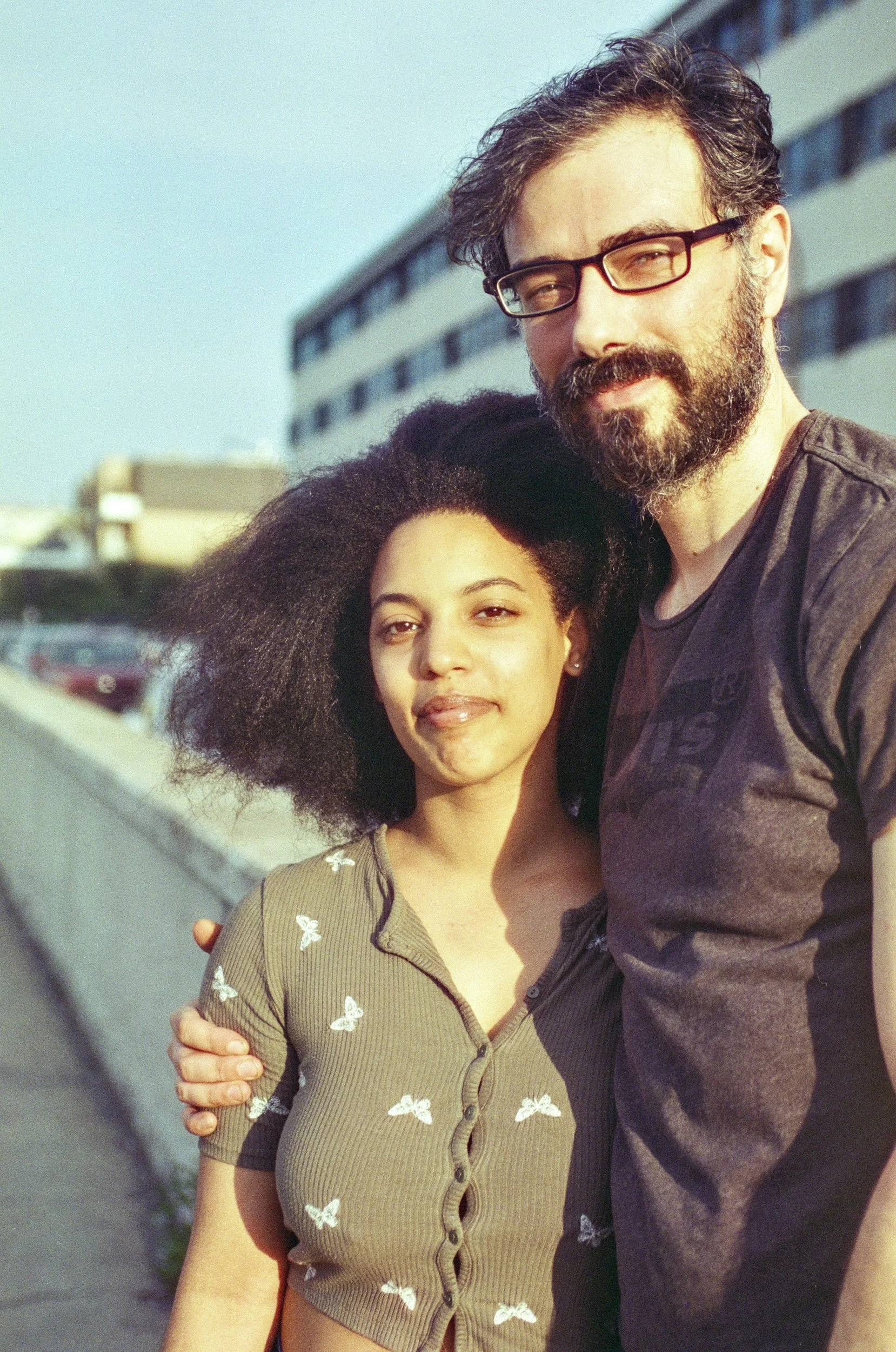 A man and a woman standing close together outside, embracing and smiling. The man has curly hair, a beard, wears glasses, and a dark shirt. The woman has dark, curly hair and is wearing a khaki-green top with butterfly patterns.