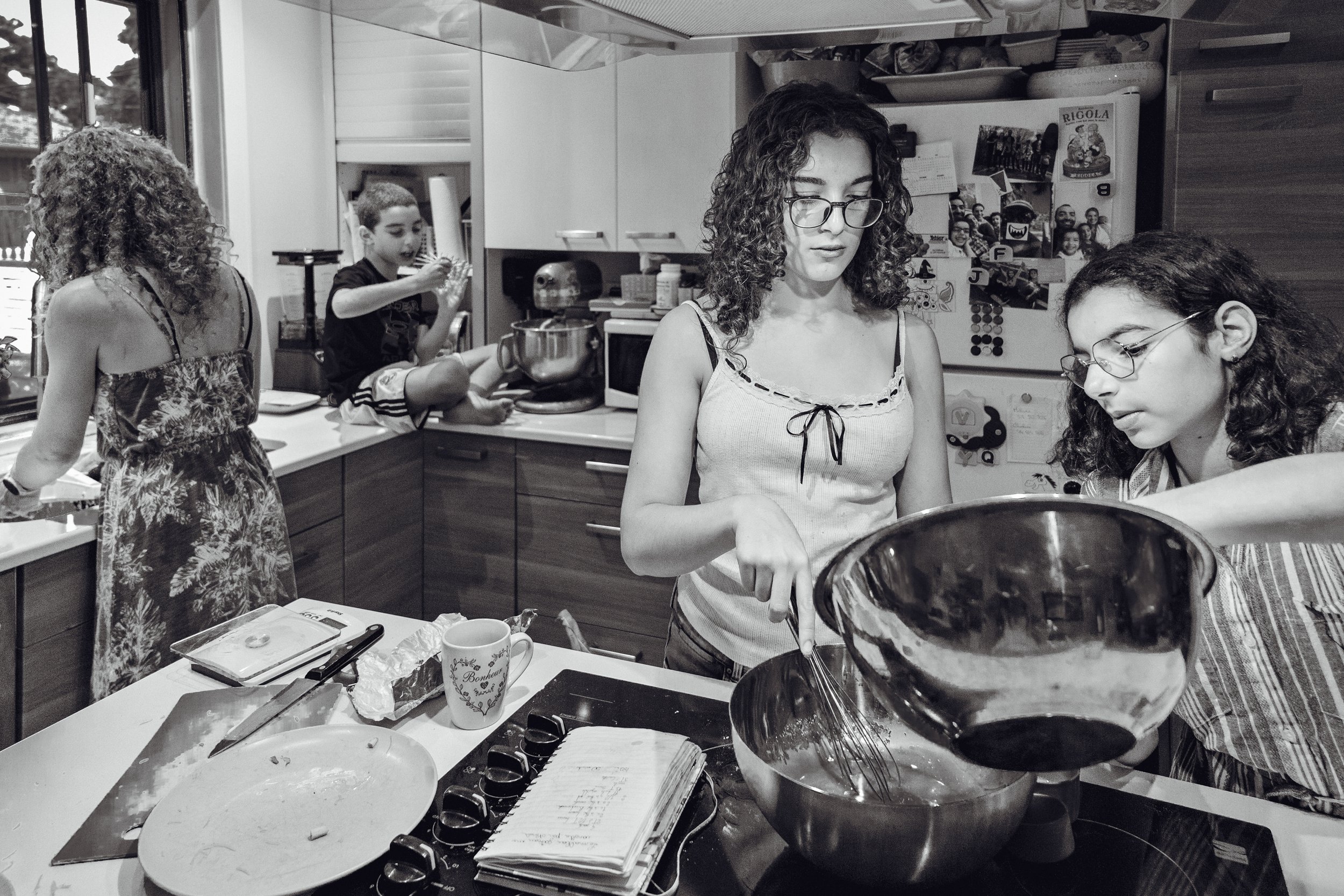 People cooking and preparing food in a kitchen, with various utensils, notebooks, and items on the counter.