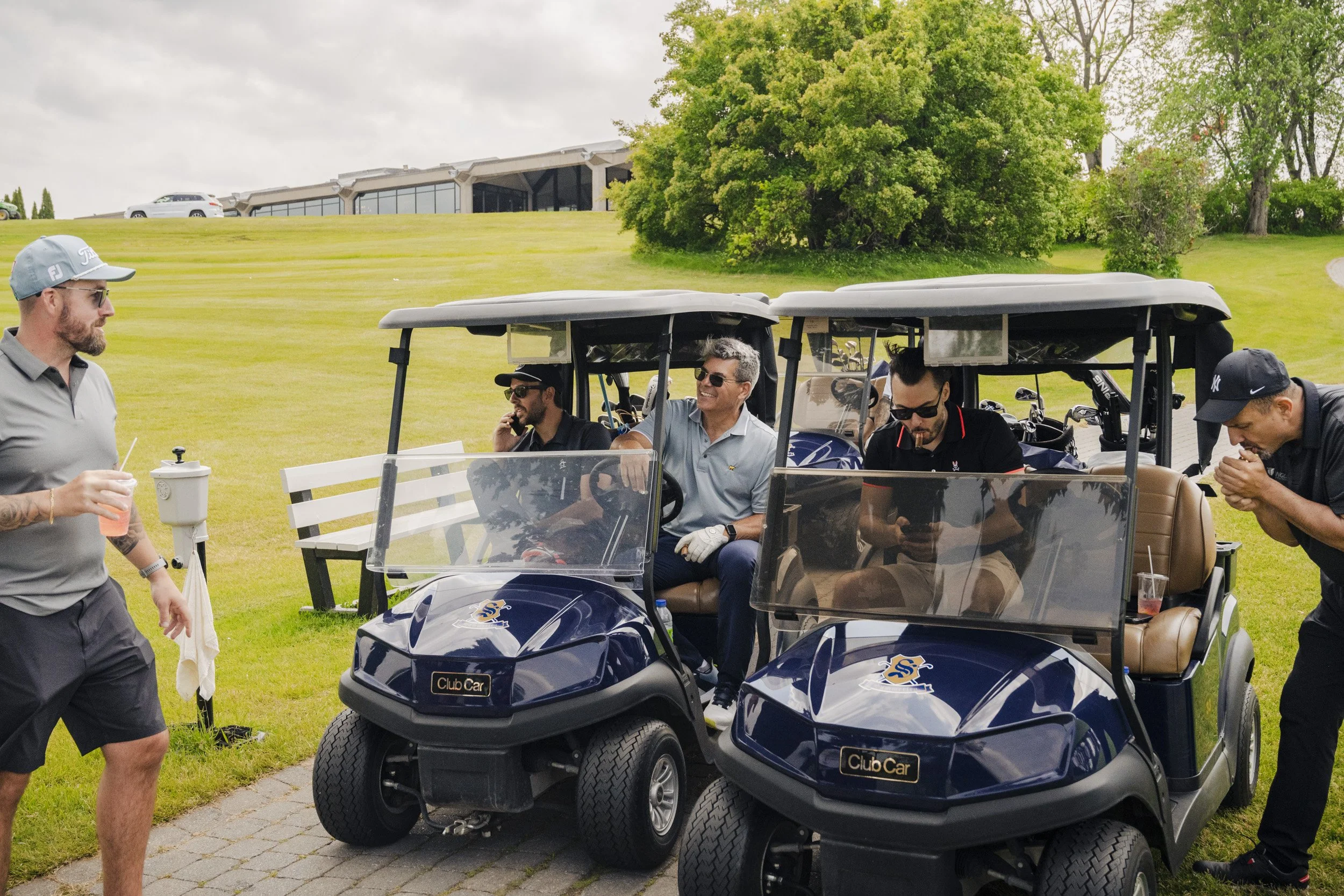 Group of men sitting and standing around blue golf carts on a golf course, with trees and a building in the background.