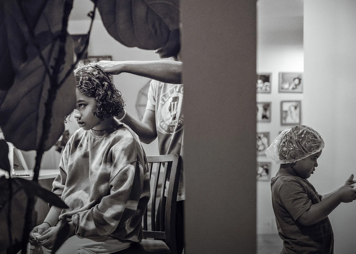 A girl with curly hair sitting in a chair at a salon or hair studio while another person styles her hair. In the foreground, a boy wearing a plastic hair cap is looking at his phone.