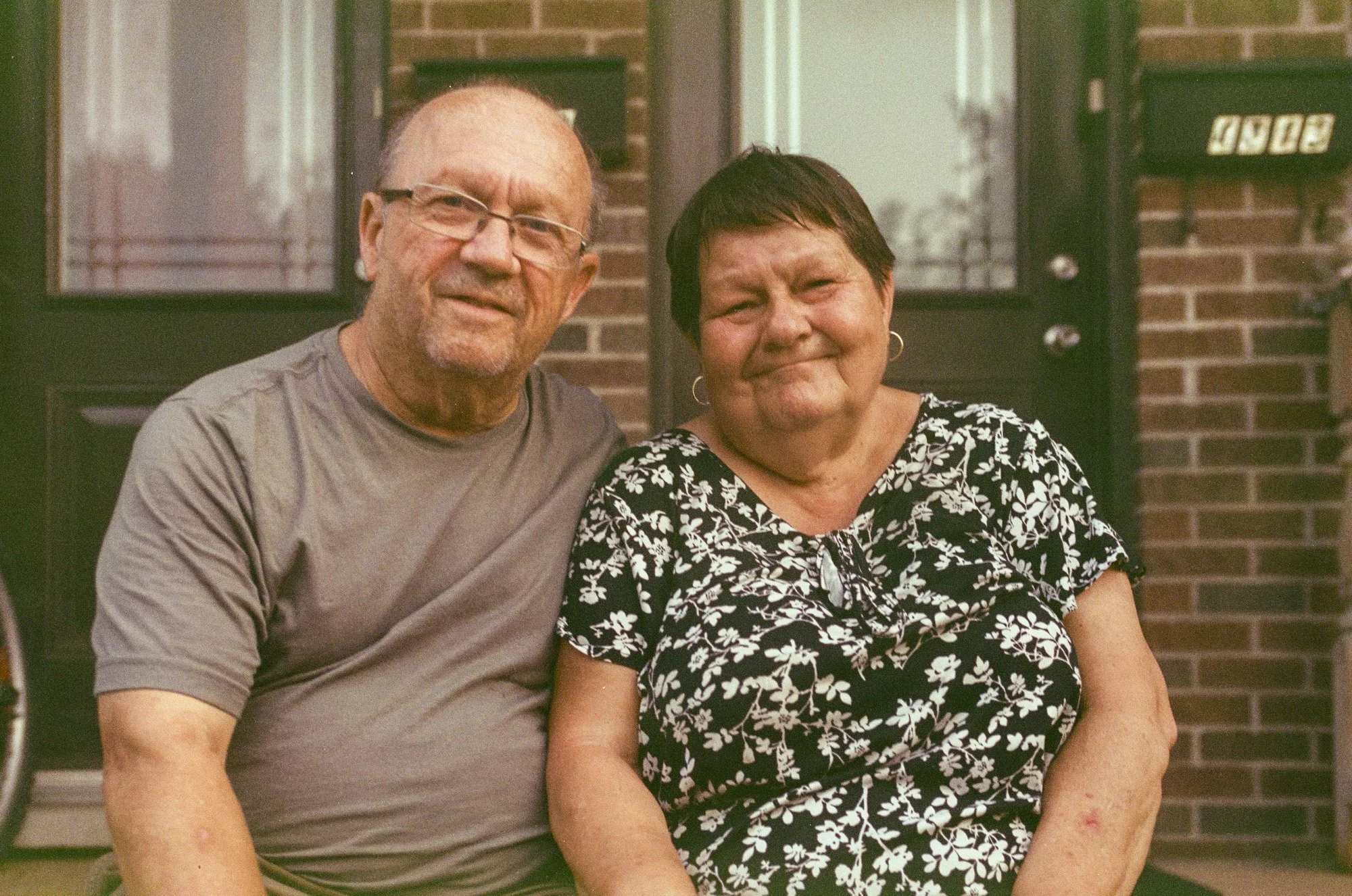 A middle-aged man with glasses and a woman with short dark hair sitting outside in front of a brick wall.