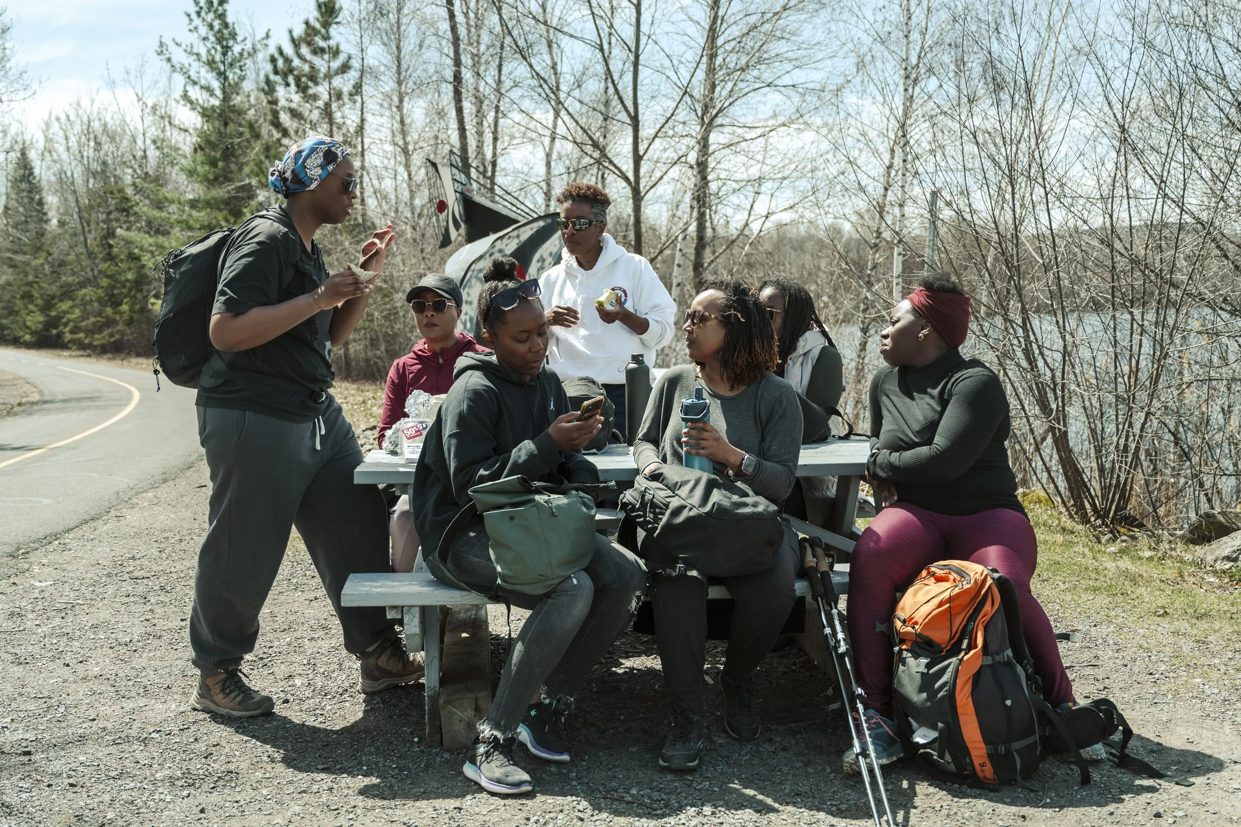 A group of women sitting at an outdoor picnic table, some wearing sunglasses, backpacks, and holding water bottles, while one woman is standing and talking to them. The scene takes place on a dirt area with trees and a helicopter in the background, i