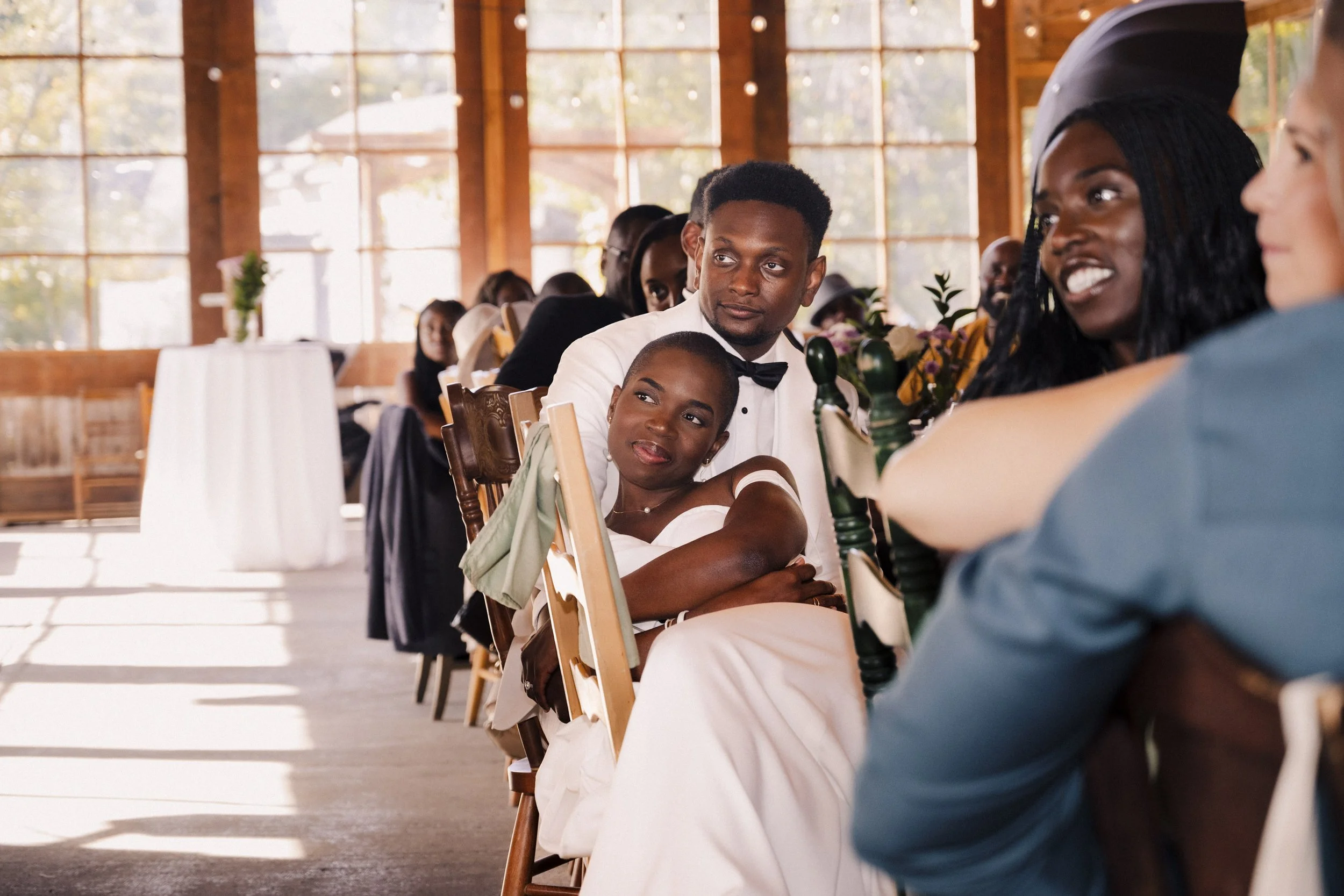 People sitting at tables in a warmly lit indoor venue with large windows, possibly a wedding or formal event.
