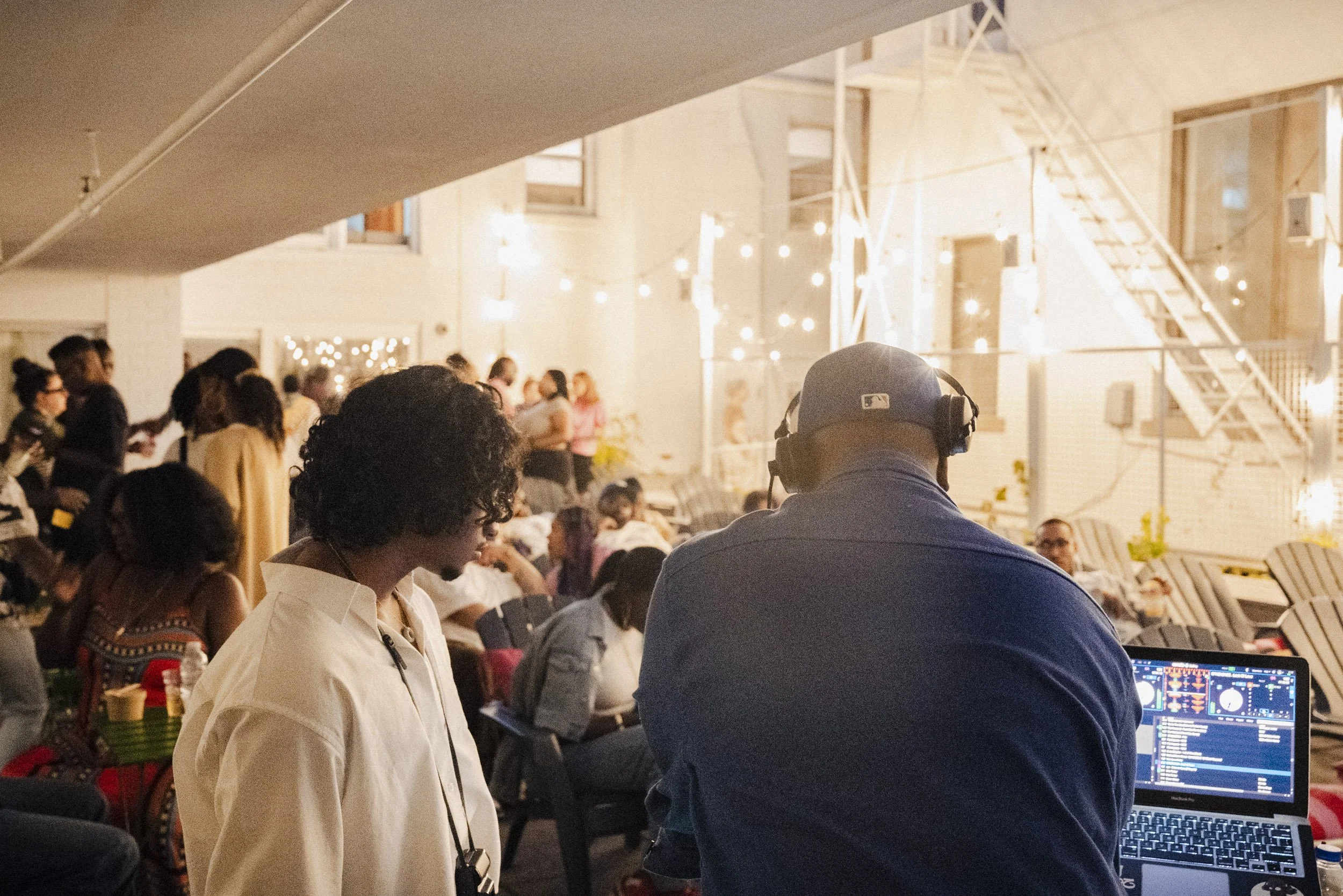 People attending an indoor event with string lights, including a man with headphones and a woman in a white shirt looking at DJ equipment.