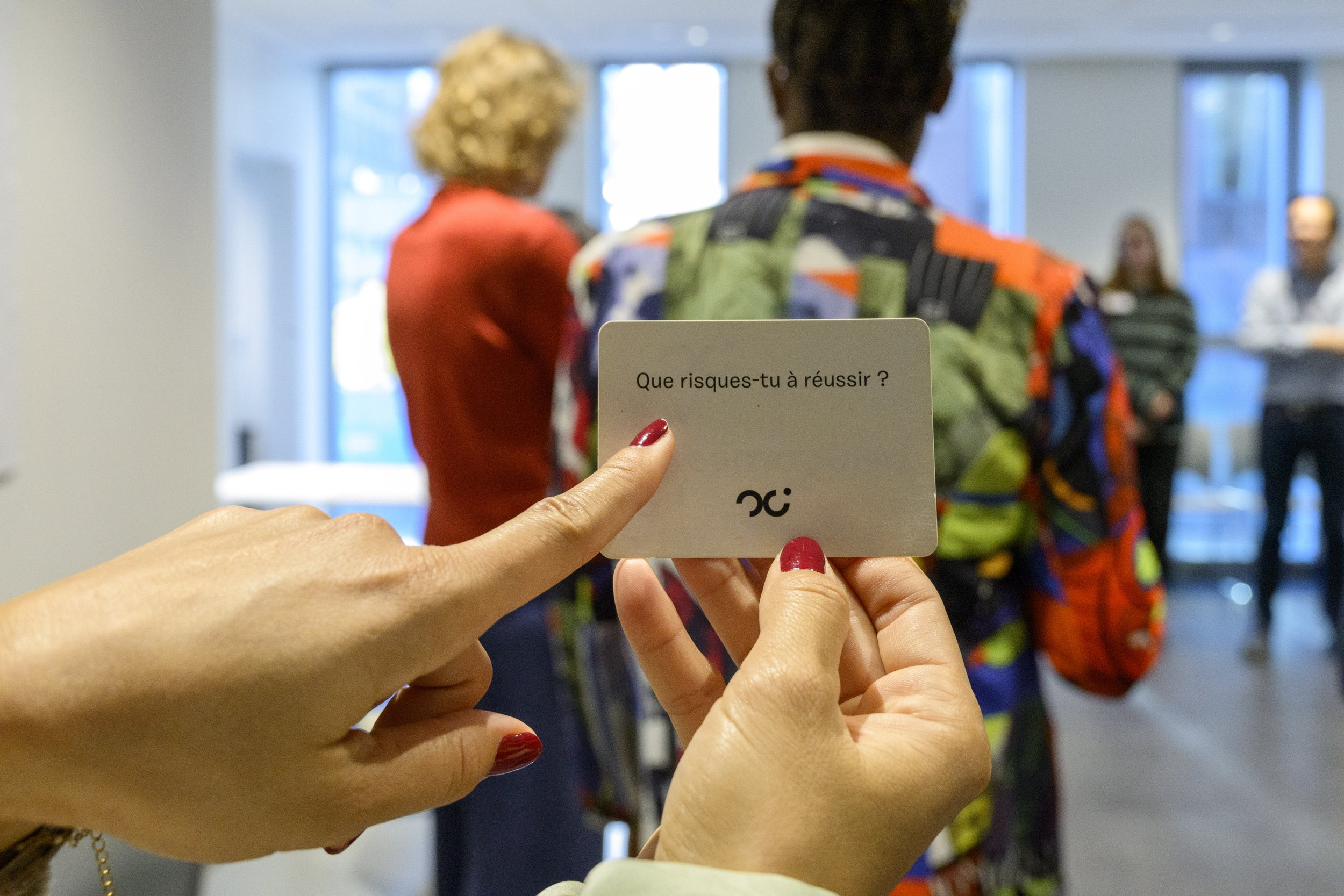 Close-up of a person's hand holding a small card with French text, 'Que risques-tu à réussir?' and a logo at the bottom. In the background are several people, some dressed in colorful clothing, in what appears to be a bright indoor setting.