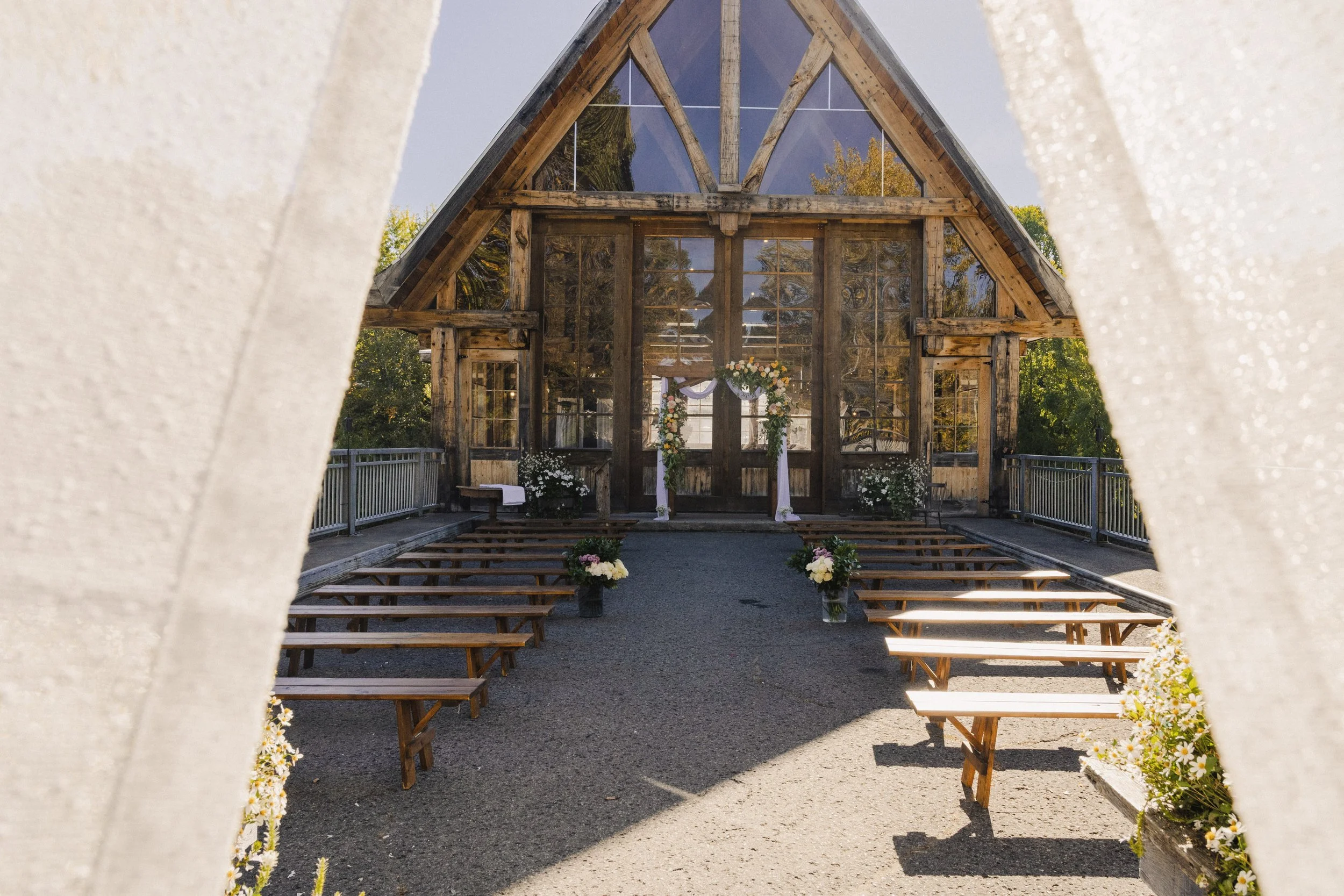 Wooden chapel or wedding venue framed by white fabric curtains with flower arrangements along the aisle.
