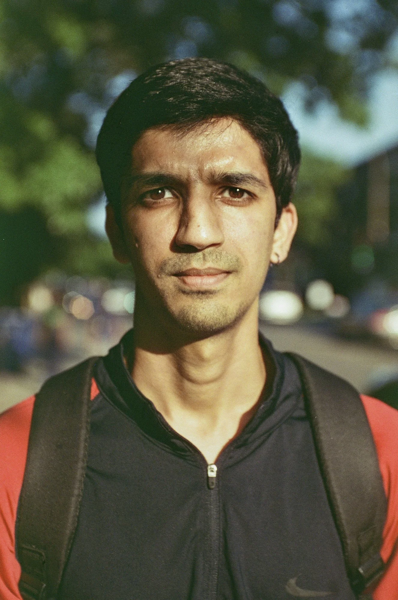 A young man with dark hair, light brown skin, and wearing a black and red sports jacket with a zipper, stands outdoors with blurred greenery and bokeh lights in the background.