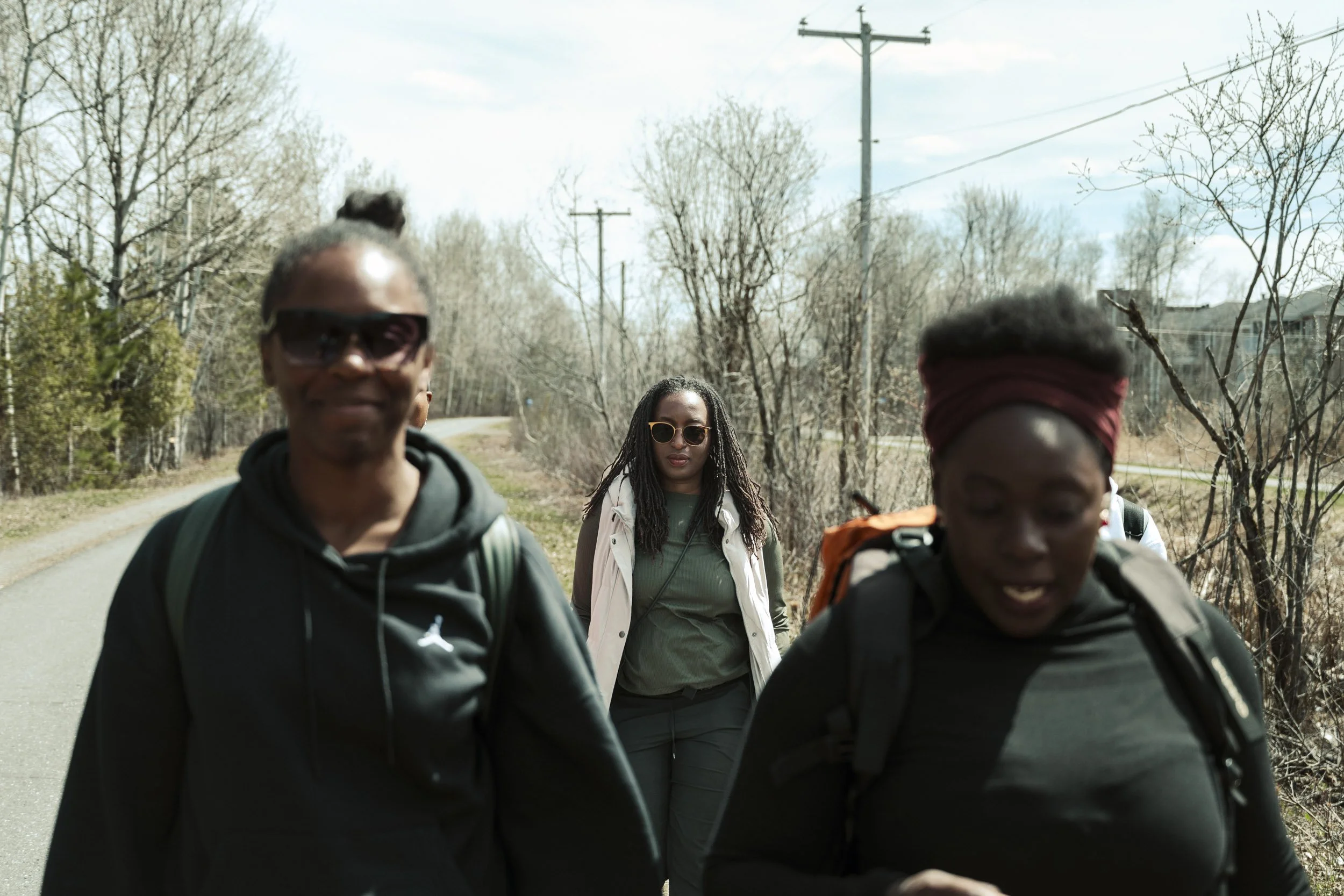 A group of young adults walking outdoors on a paved path during daytime, surrounded by leafless trees and utility poles, dressed casually with backpacks and sunglasses.