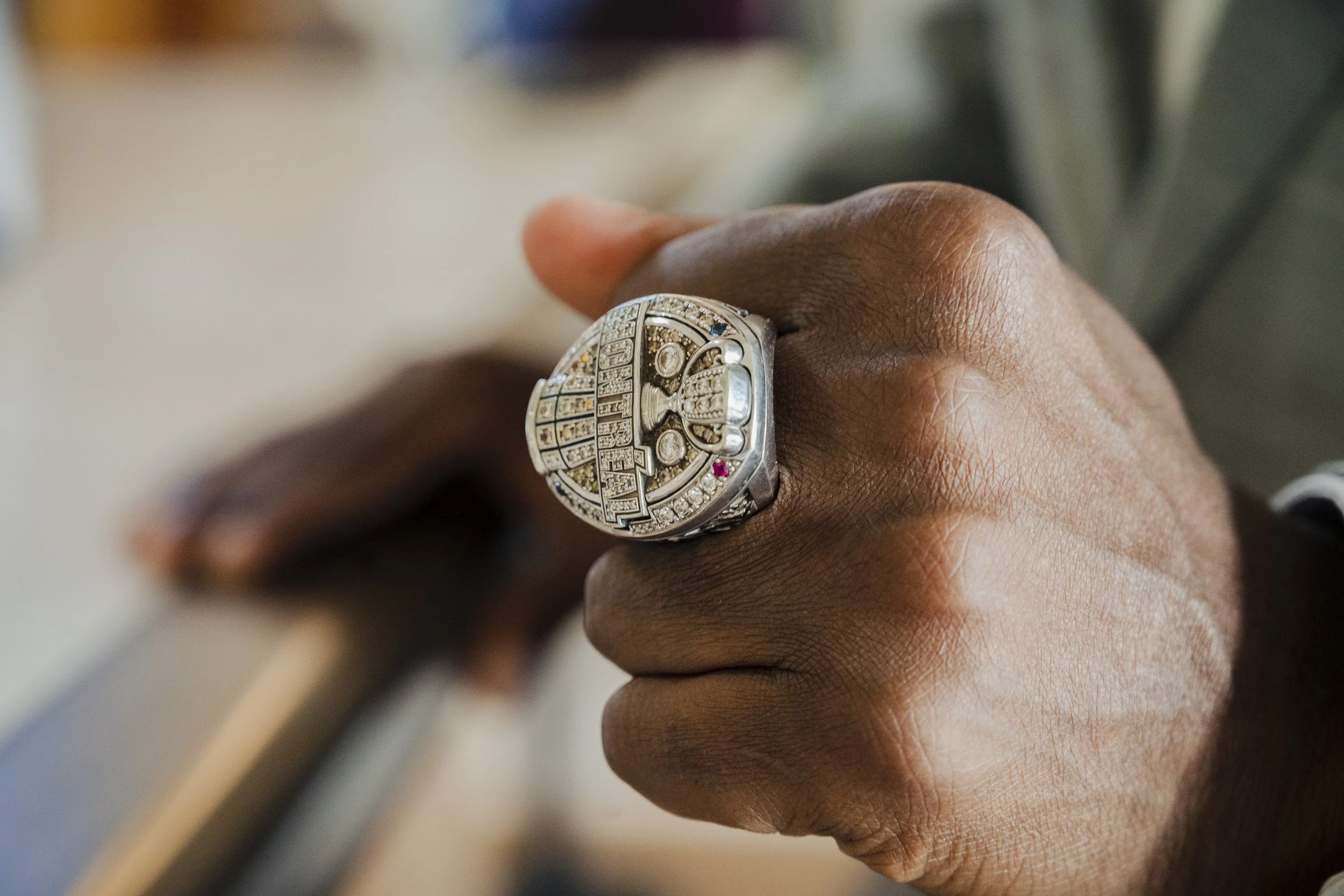 Close-up of a person's hand wearing a championship ring with diamonds and text, against a blurred background.