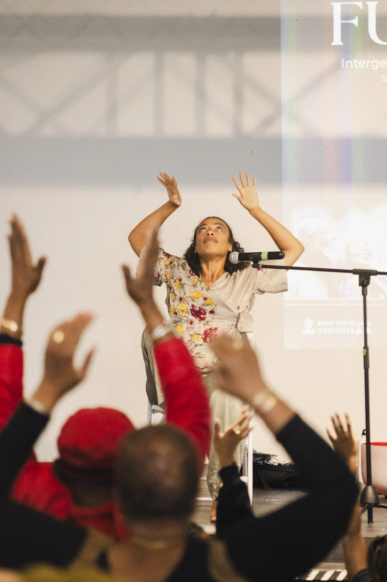 A woman with dark, curly hair is on stage with a microphone, raising her arms above her head, performing in front of an audience. The audience, partially visible in the foreground, includes children with raised hands, some wearing red clothing. There