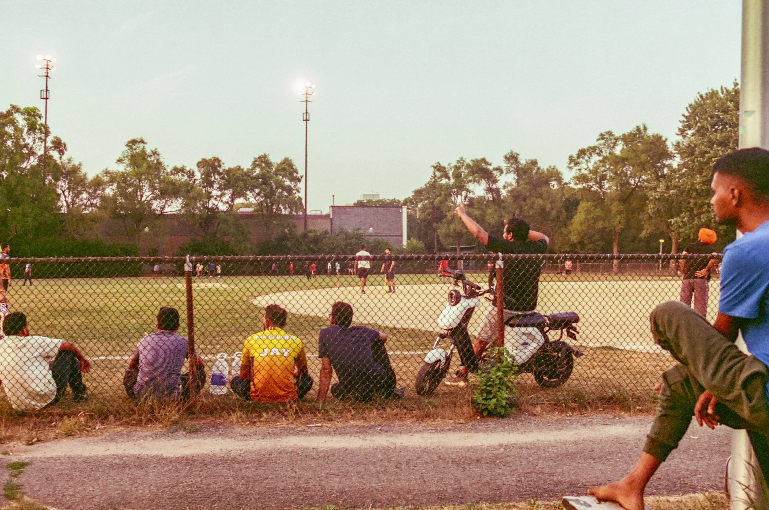 People gathered at a baseball field during sunset, some sitting and some standing, with a person in a black shirt throwing a ball; chain-link fence in front, trees and sky in the background.