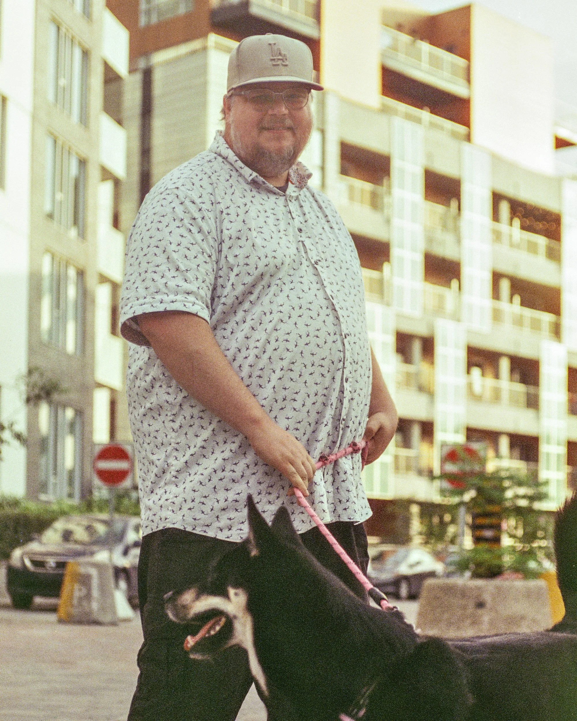 A man wearing a gray baseball cap, glasses, and a white shirt with small black patterns stands outside near modern apartment buildings. He is holding a pink leash attached to a black dog with white markings.