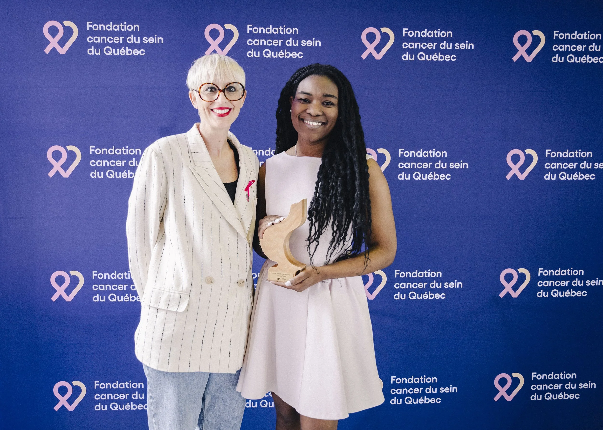 Two women standing in front of a blue backdrop with pink ribbon logos and text that reads 'Fondation cancer du sein du Québec'. One woman has short, blonde hair, glasses, and is wearing a striped blazer. The other woman has long, dark, braided hair a
