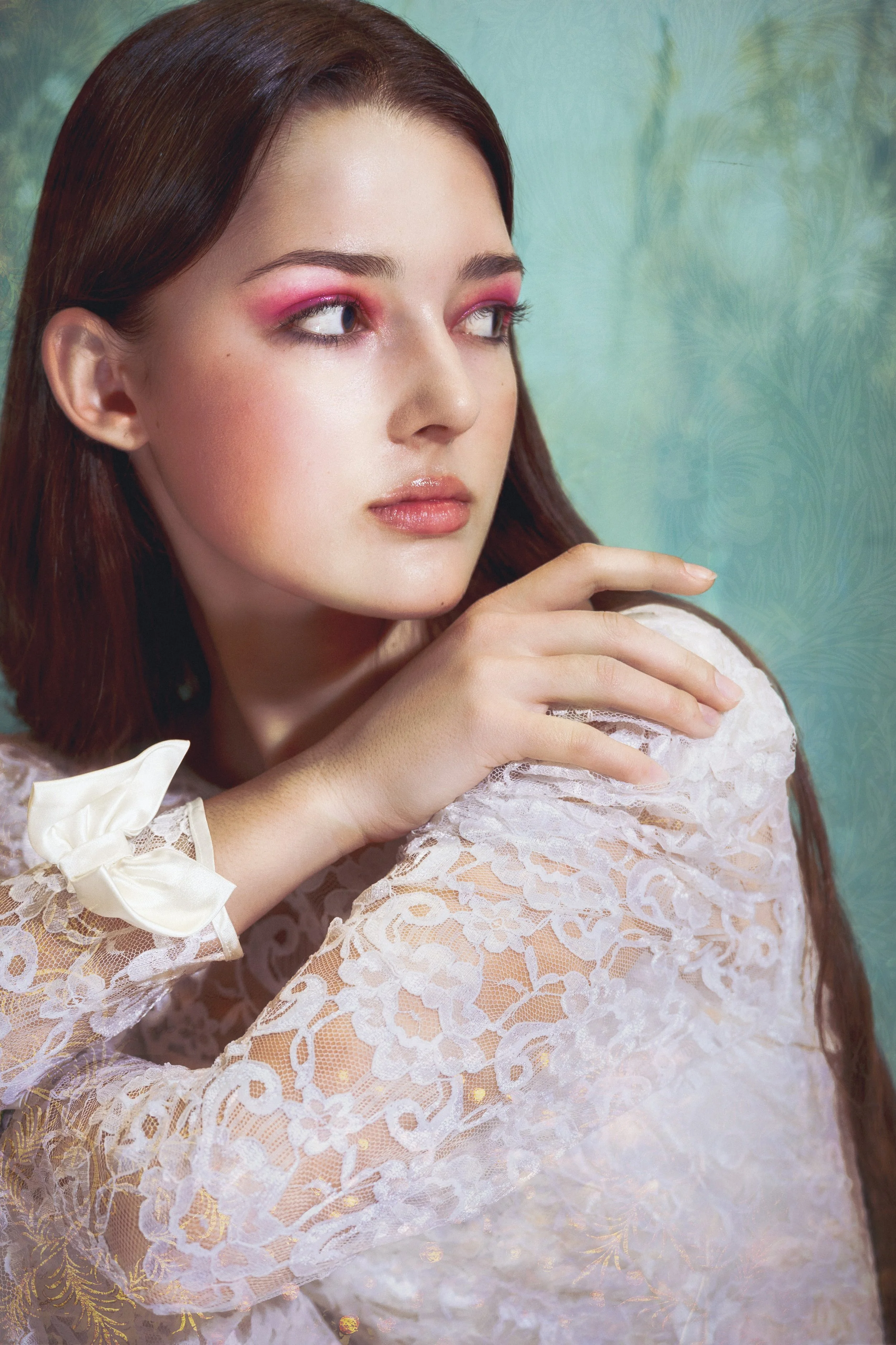 A young woman with long red hair and bright makeup, wearing a white lace dress with a bow on the sleeve, standing against a teal background with a floral pattern.