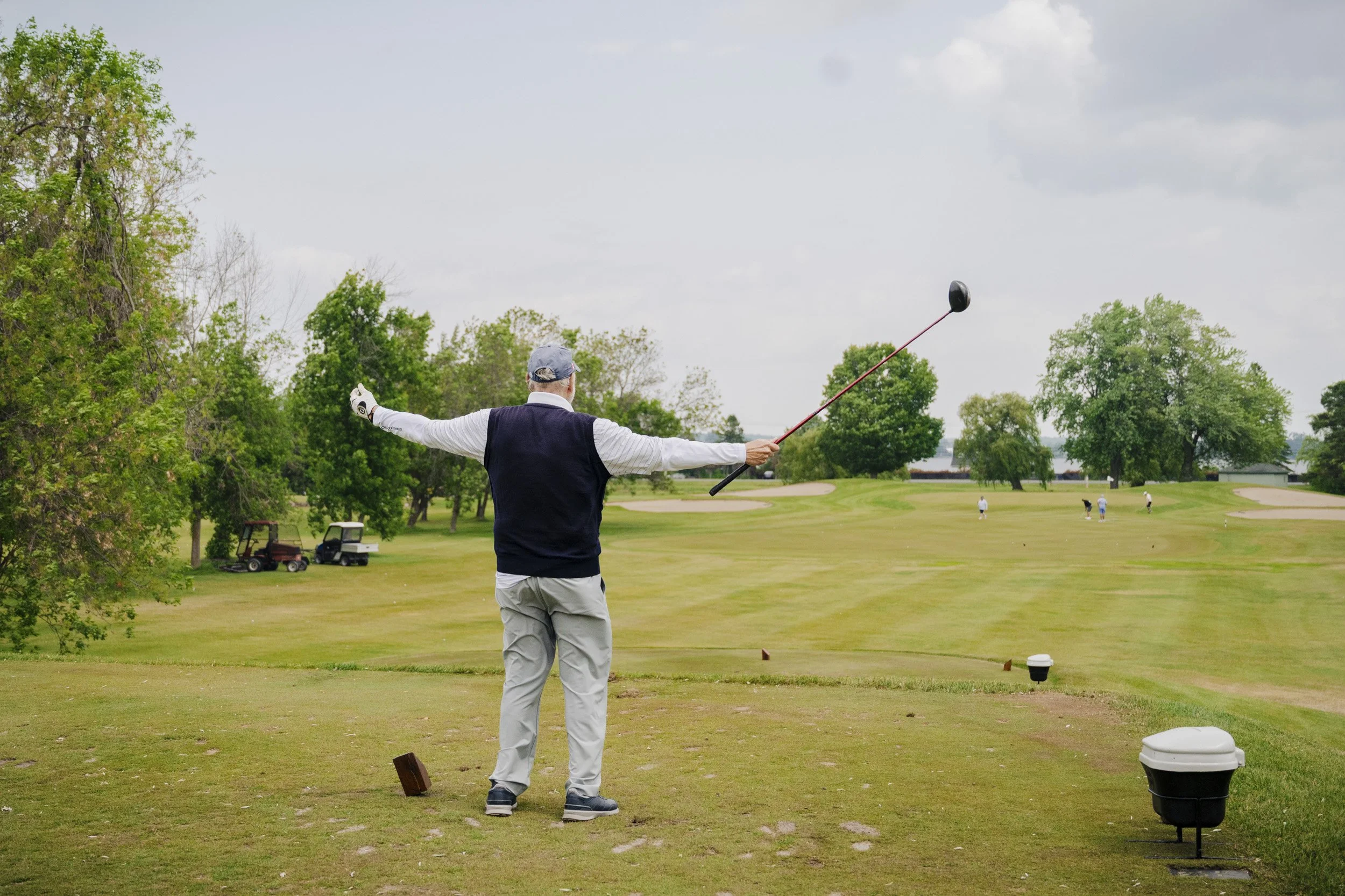 A man on a golf course preparing to hit a golf ball, dressed in a golf cap, a polo shirt, and light-colored pants, surrounded by lush trees and open sky.