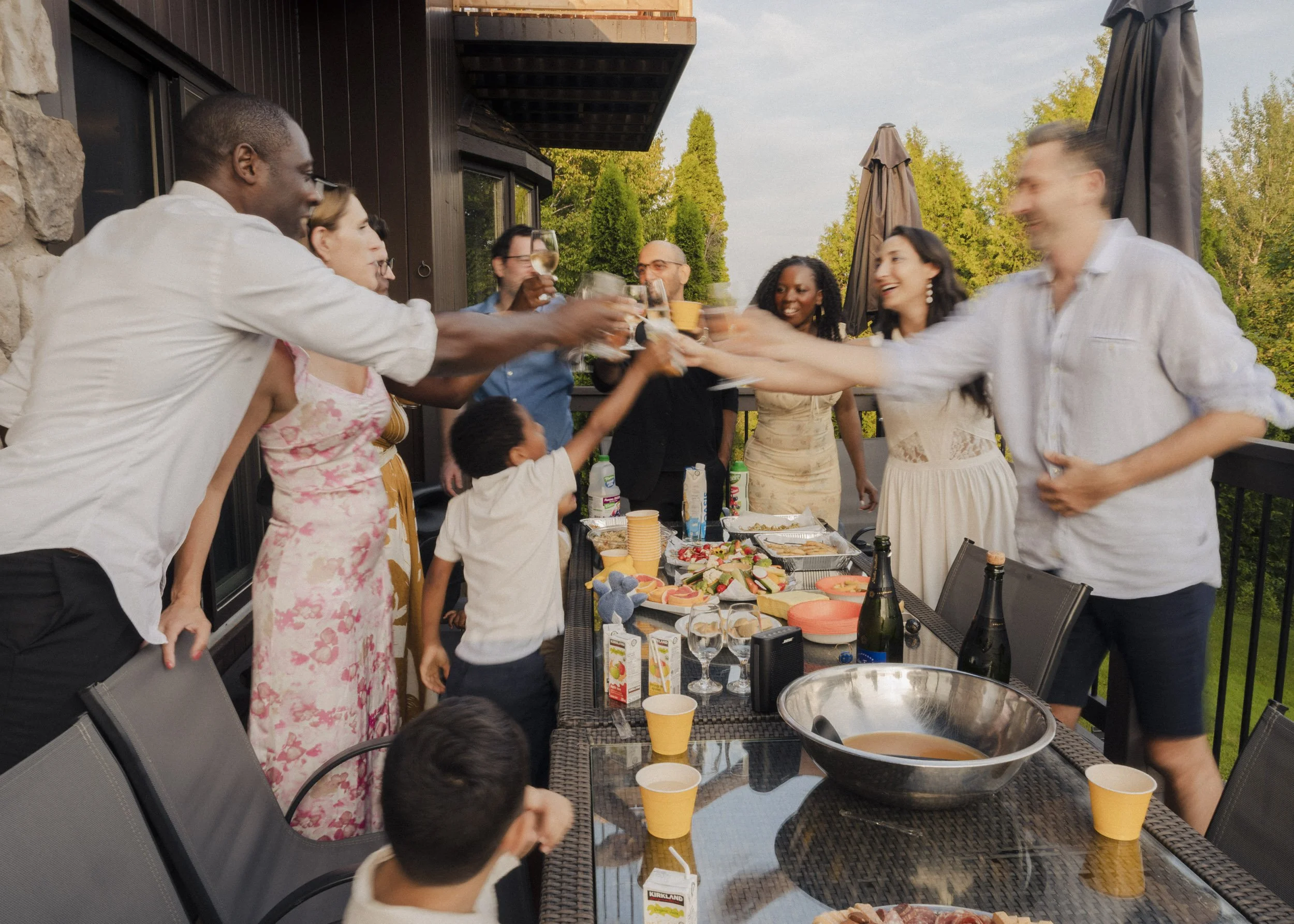 A group of people at an outdoor party, toasting glasses over a table with food, drinks, and snacks on a patio with trees in the background.