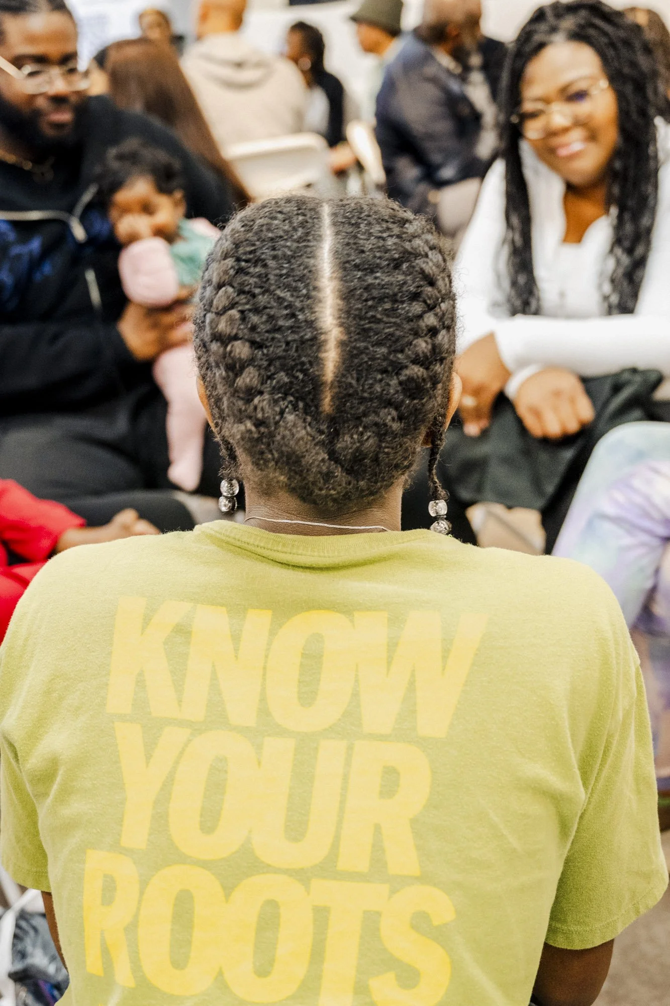 Back of a person wearing a yellow t-shirt with large yellow text reading 'KNOW YOUR ROOTS' sitting in a crowded indoor setting with people talking and a woman smiling with glasses and long hair.