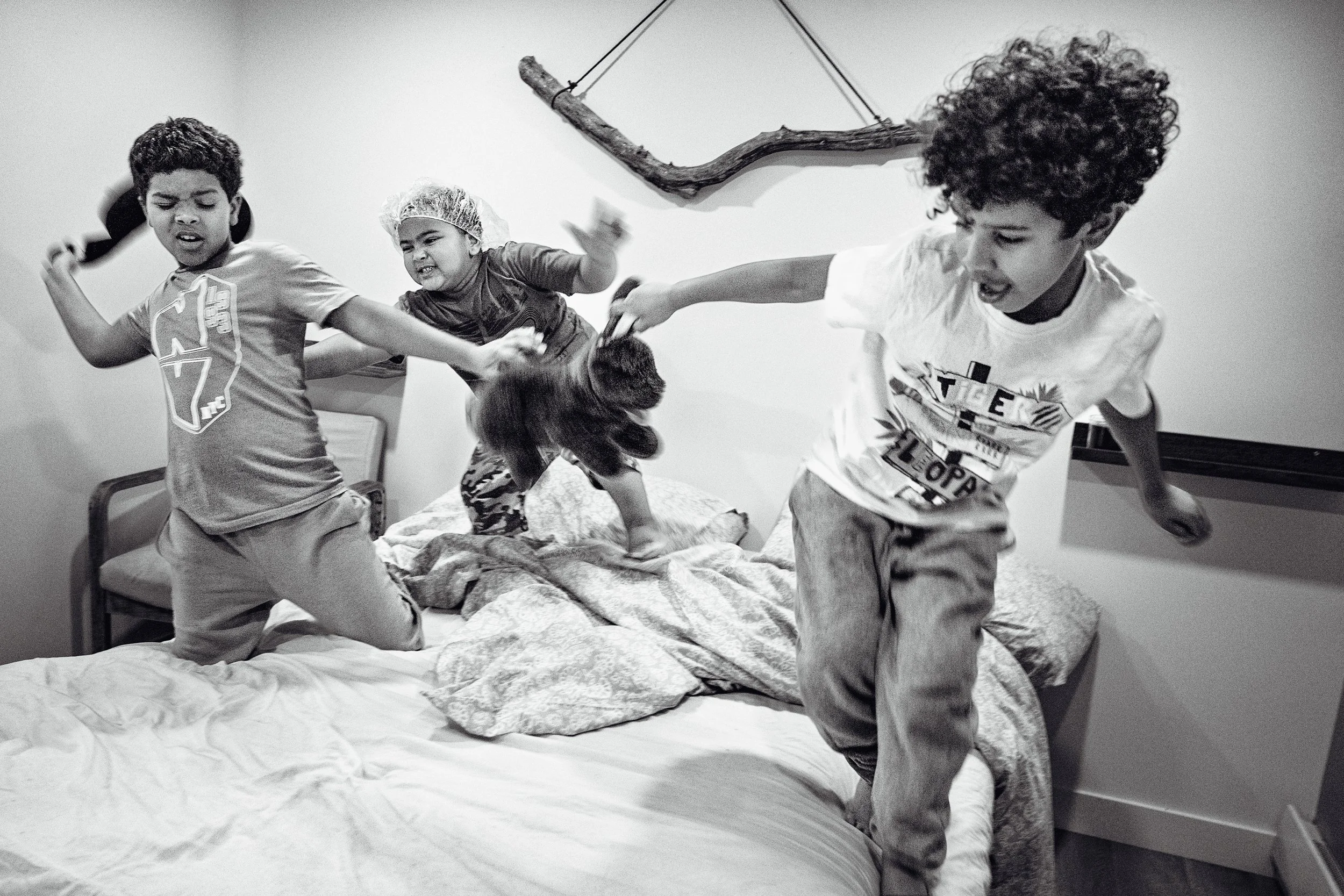 Three children playing and fighting on a bed in a bedroom. Two boys and a girl, one boy has curly hair and the girl is wearing a shower cap, all appearing to be having fun or playing roughly.