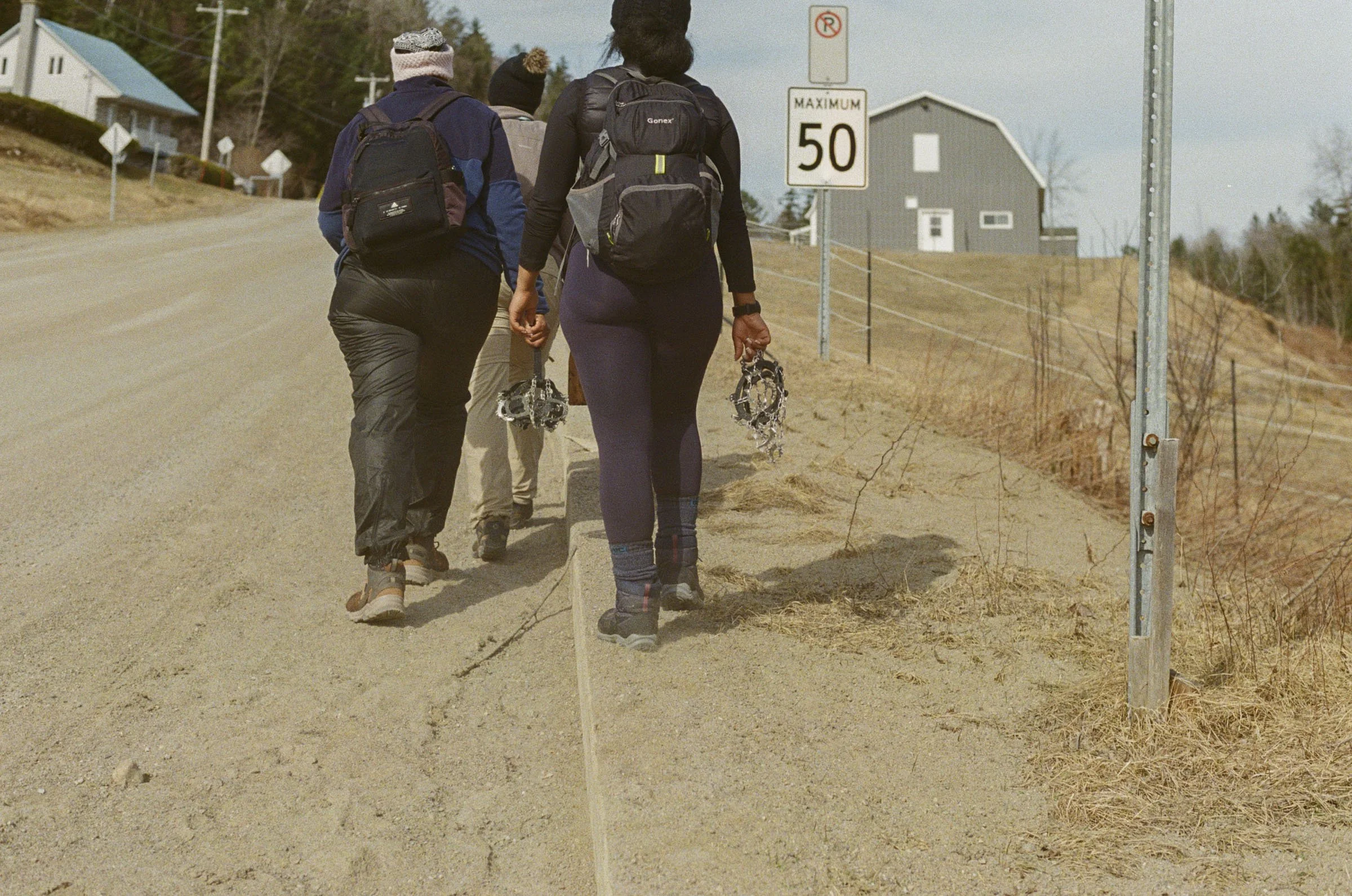 Three people walking on the side of an empty road with backpacks, holding snowshoes, in a town with houses, a street sign showing a 50 mph speed limit, and dry grass on the roadside.