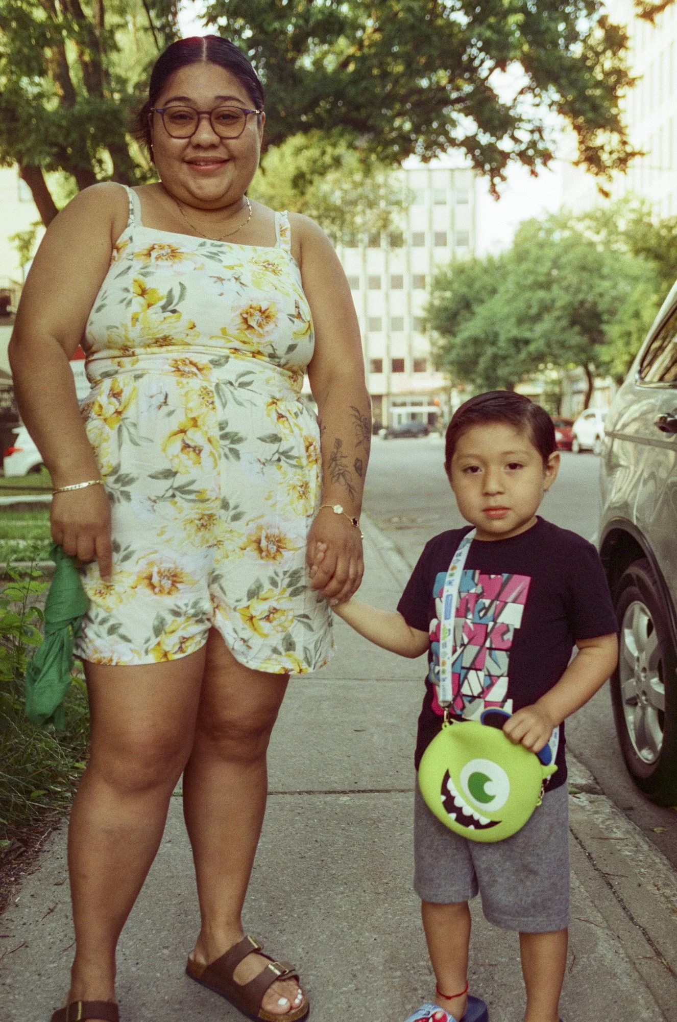 A woman and a young boy standing outdoors on a sidewalk, holding hands. The woman is wearing glasses, a white floral tank top, and shorts. The boy is wearing a black graphic T-shirt, gray shorts, and carrying a green monster-themed lunchbox. They are