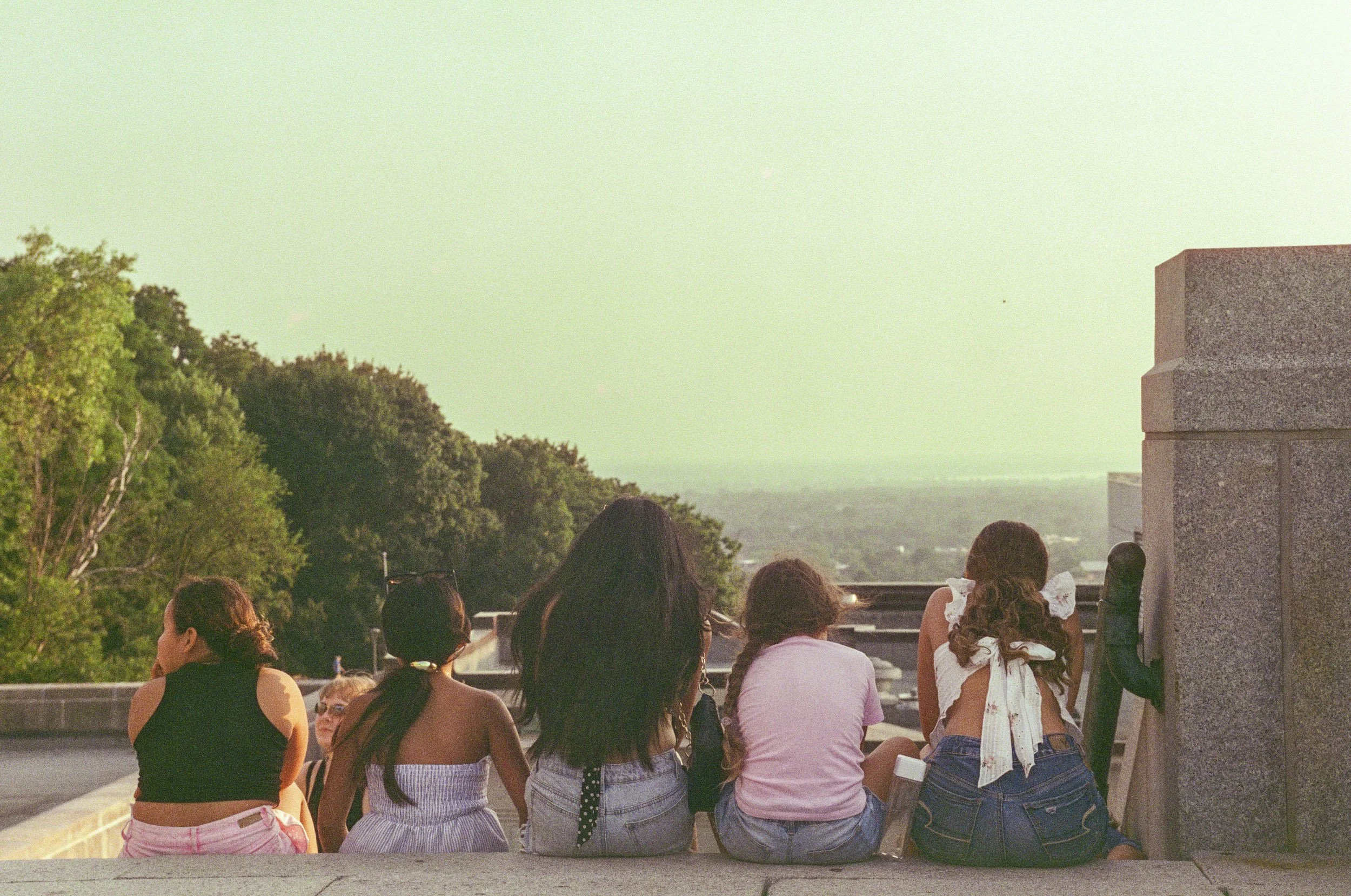 Group of young women sitting on a ledge outdoors, facing away, with trees and a distant landscape in the background during daylight.