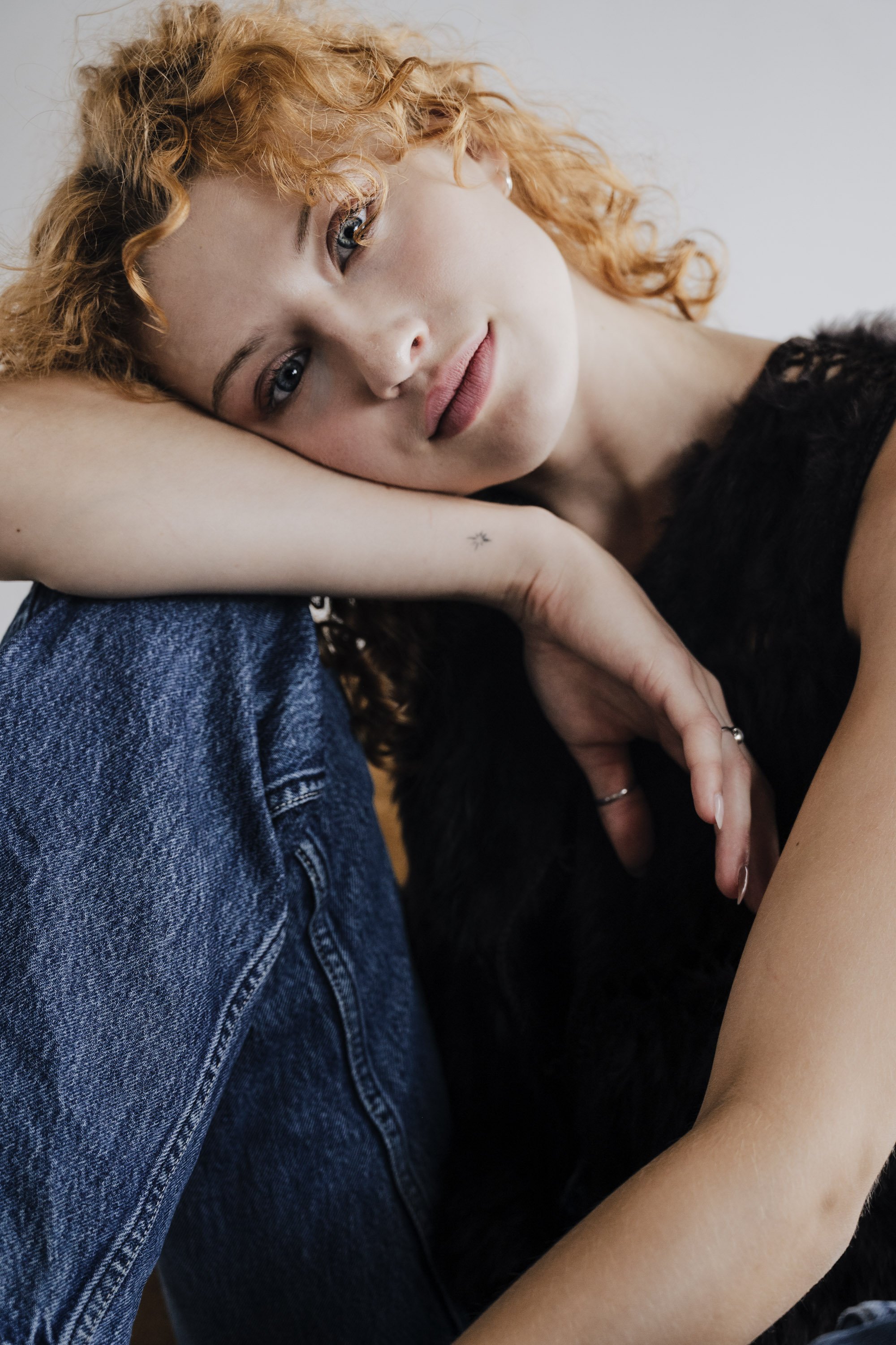 Close-up of a young woman with curly red hair lying on her side, resting her head on her arm, and looking at the camera with a gentle expression.