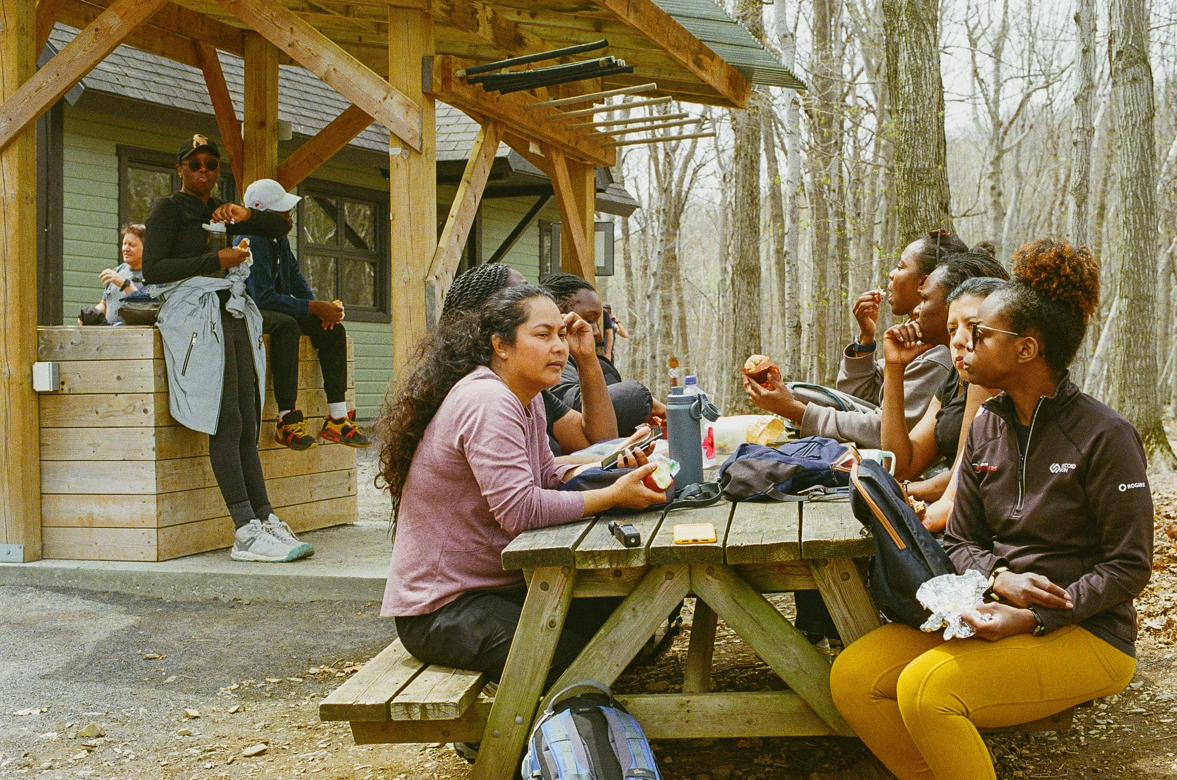 Group of people sitting and standing around an outdoor wooden picnic table and bench in a wooded park, eating snacks and engaging in conversation during daytime.
