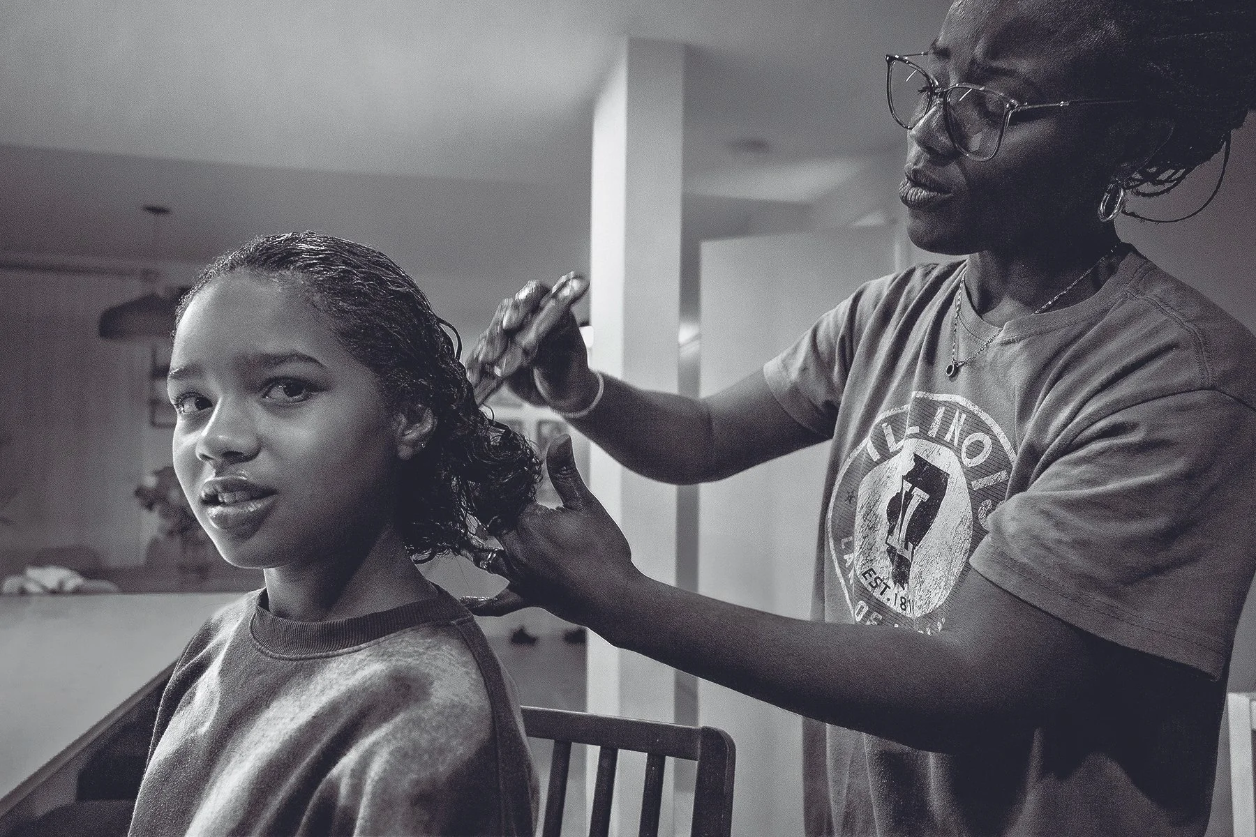 A woman with curly hair getting her hair styled by another woman wearing glasses, in a black-and-white photo.