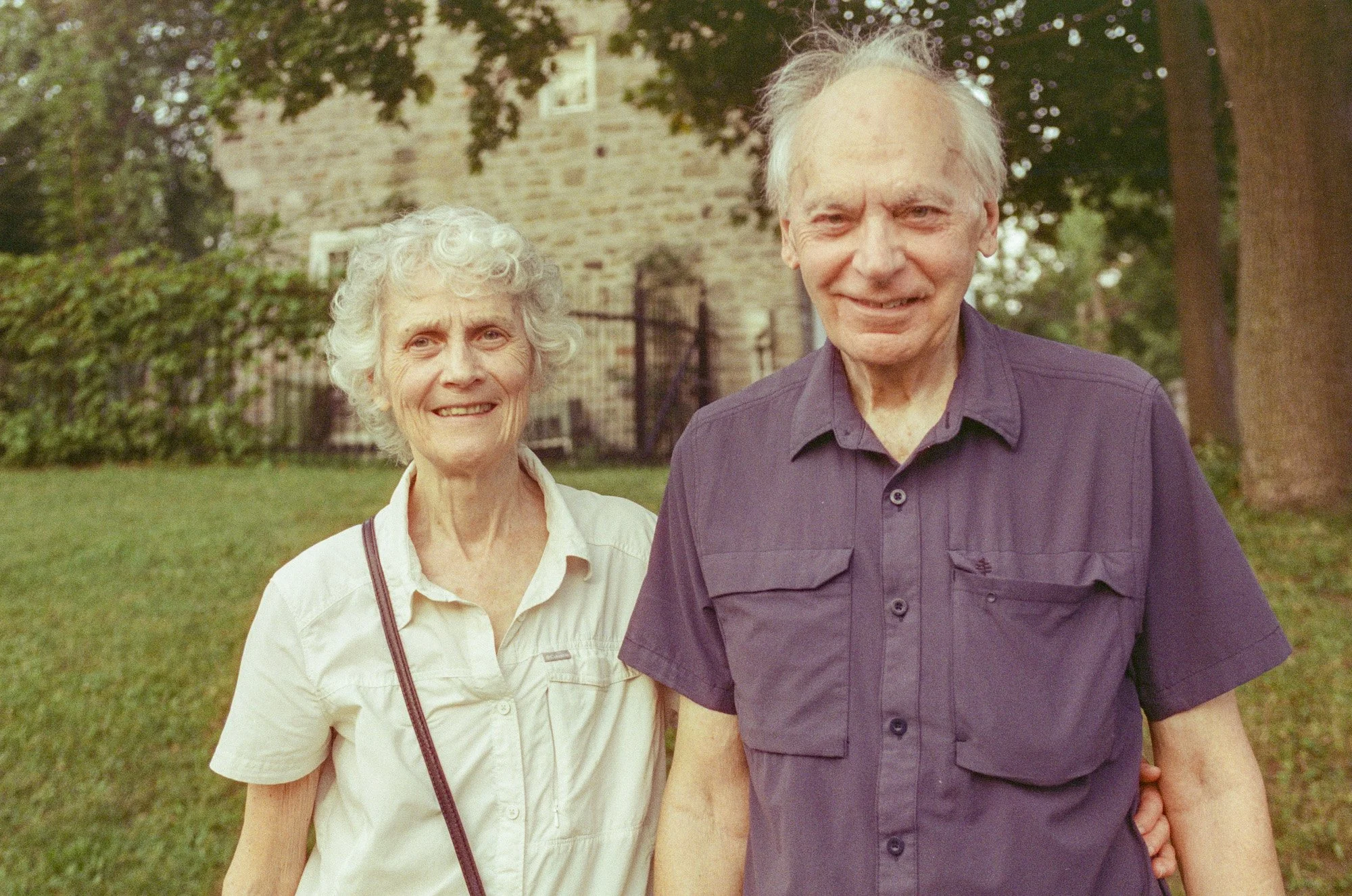 An elderly woman and man smiling outdoors, standing on grass with trees and a brick building behind them.