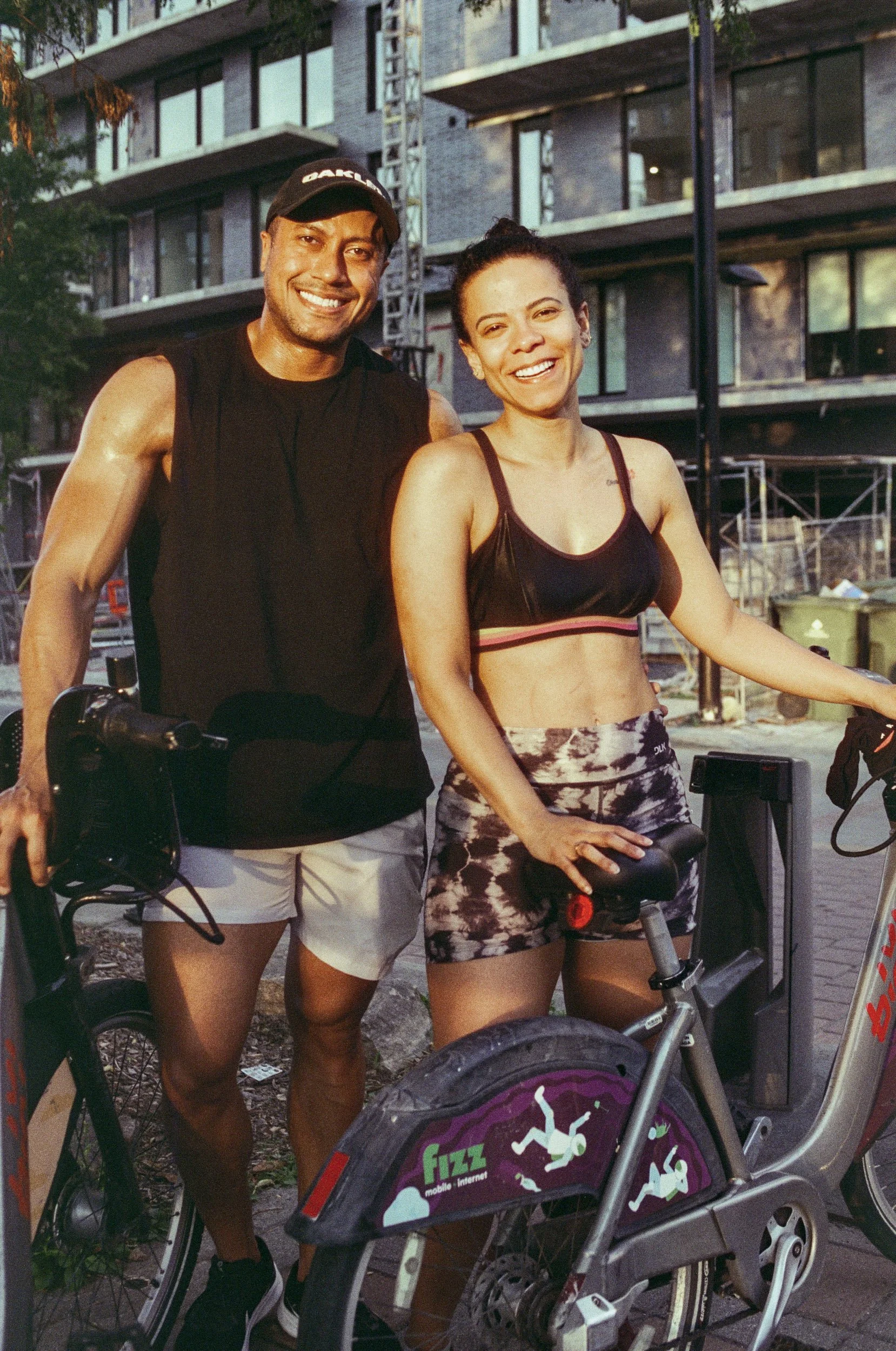 A smiling man and woman standing outdoors next to a bicycle, with a modern building in the background.