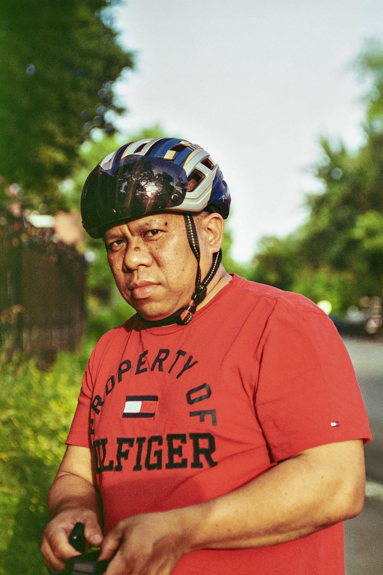 Man wearing a black helmet with a reflective visor, red T-shirt with text, outdoors with blurred green trees and sky in background.