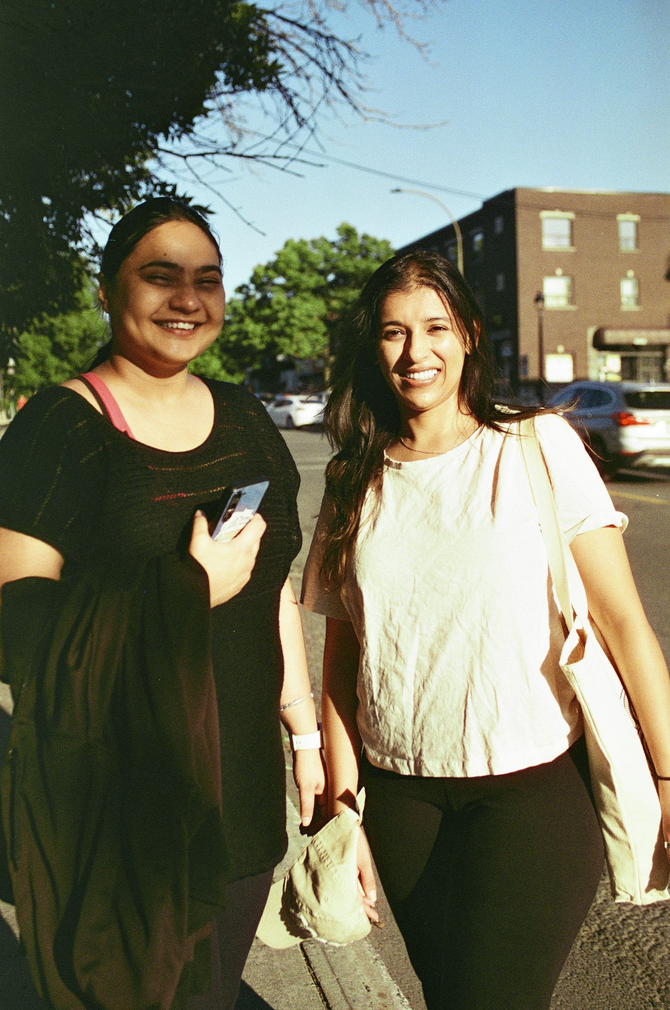 Two smiling women standing outdoors on a sunny day, with cars and a building in the background.