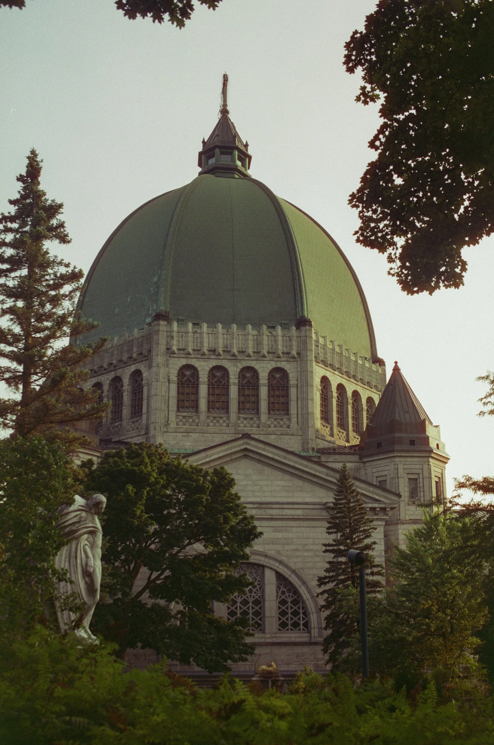A historic building with a large green dome and a spire, surrounded by trees and a statue of a person in the foreground.
