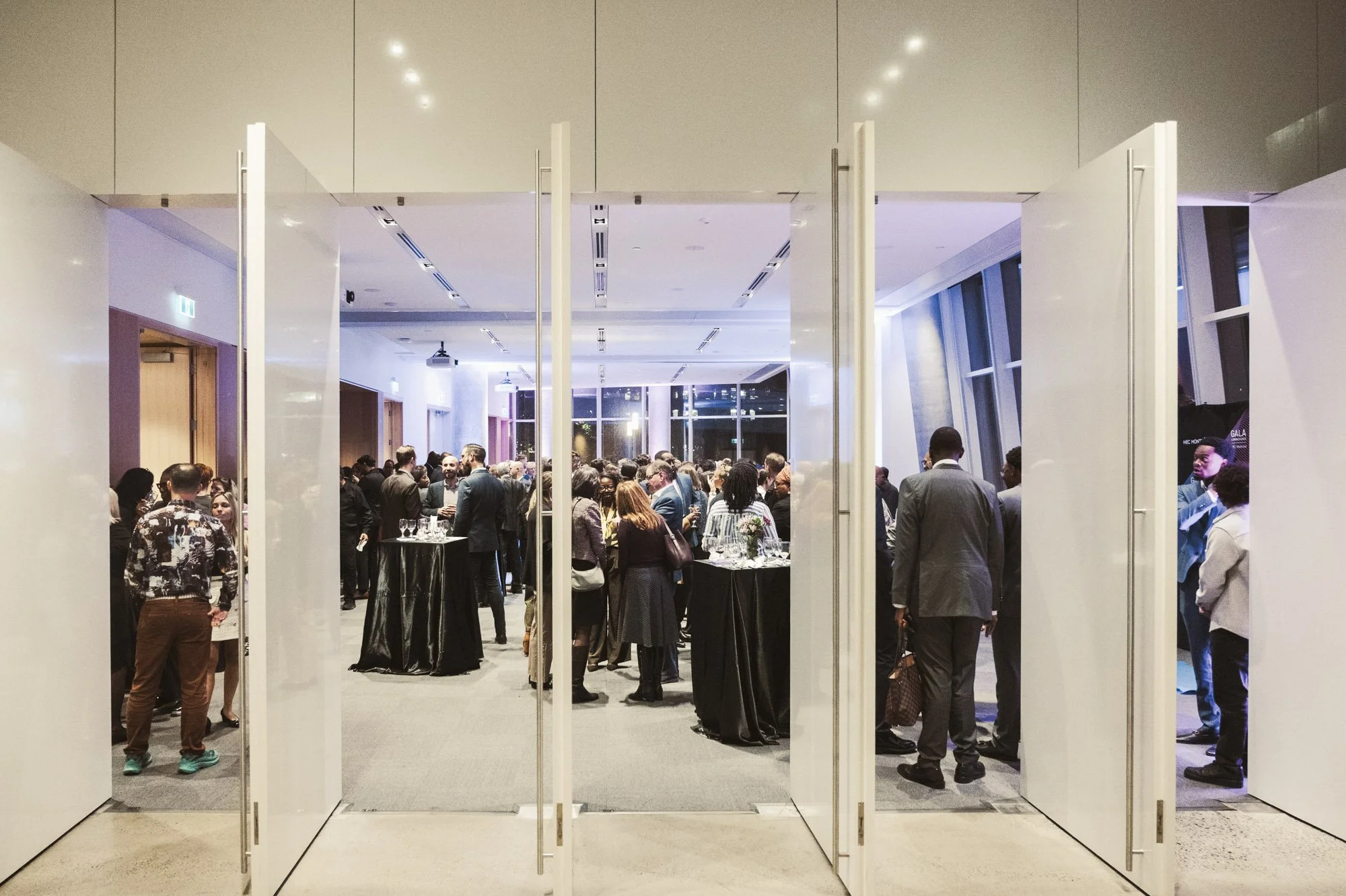 A large social gathering in a modern, well-lit interior space viewed through open doors. People are mingling, talking, and standing around tables, with some wearing business attire. The room features large windows and a high ceiling.