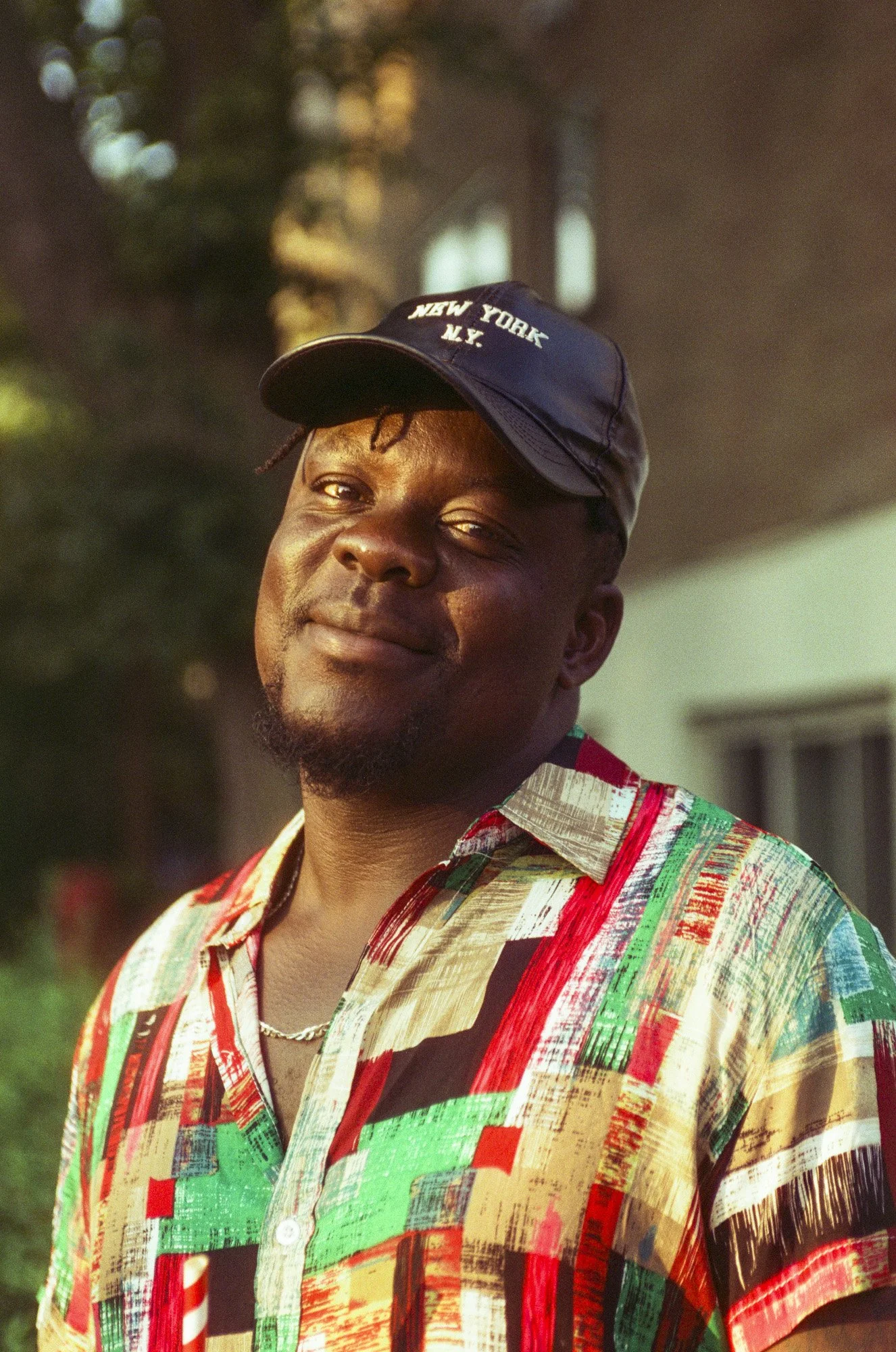 A man wearing a colorful, patterned shirt and a black baseball cap with 'New York N.Y.' embroidered on it, standing outdoors during the daytime.