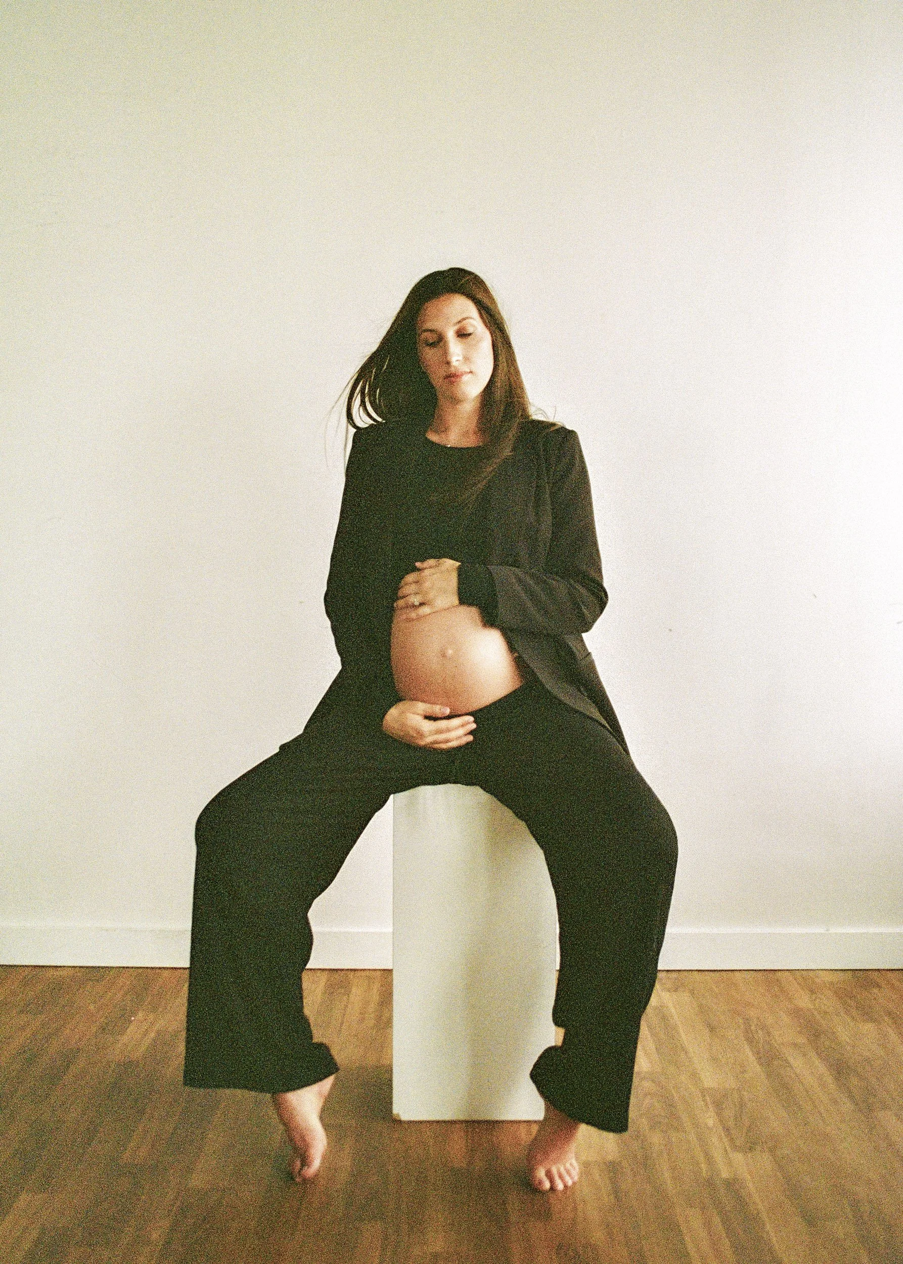 A woman in black pants and a black blazer is standing with her legs wide apart, arms crossed in front of her pregnant belly, in front of a white wall and wooden floor.