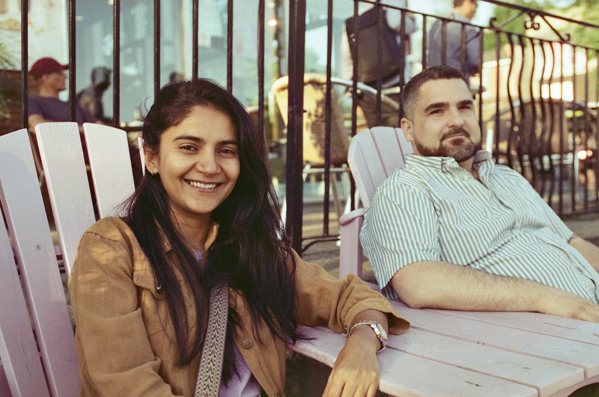 Two people sitting on outdoor chairs, smiling and relaxing on a sunny day. The woman has long dark hair, is wearing a brown jacket, and appears happy. The man has short dark hair, a beard, and is wearing a striped shirt, looking relaxed.