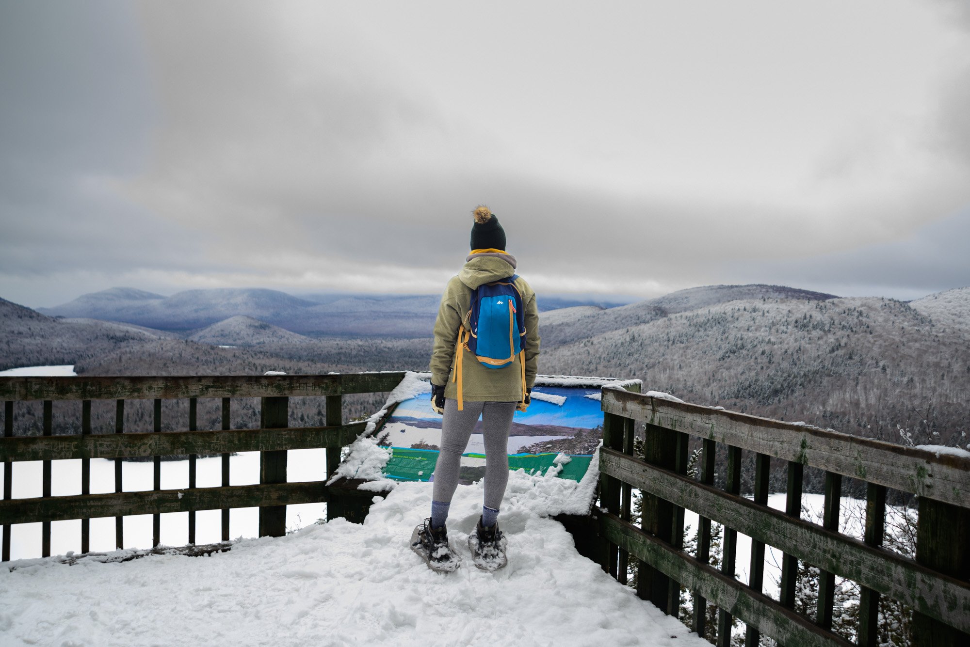 Person in winter clothing, including a beanie, standing on a snow-covered observation deck, looking at a foggy mountain landscape with snow-covered hills and trees.