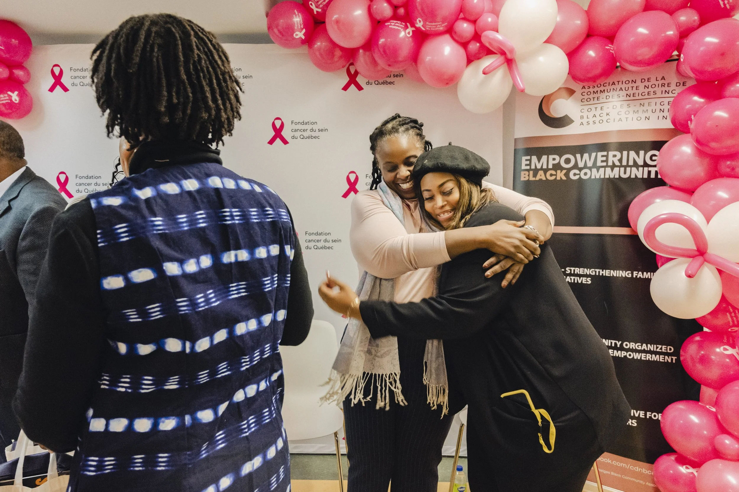 Two women hugging each other at an awareness event with pink and white balloons and a backdrop with pink ribbons and text about breast cancer awareness.