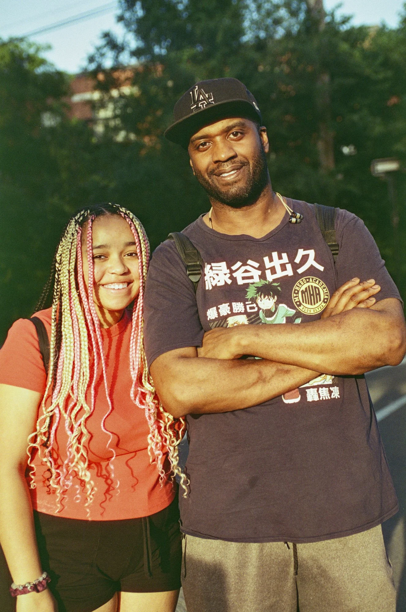 A young woman with long braided hair smiling and standing next to a man with crossed arms, both outdoors with trees in the background.