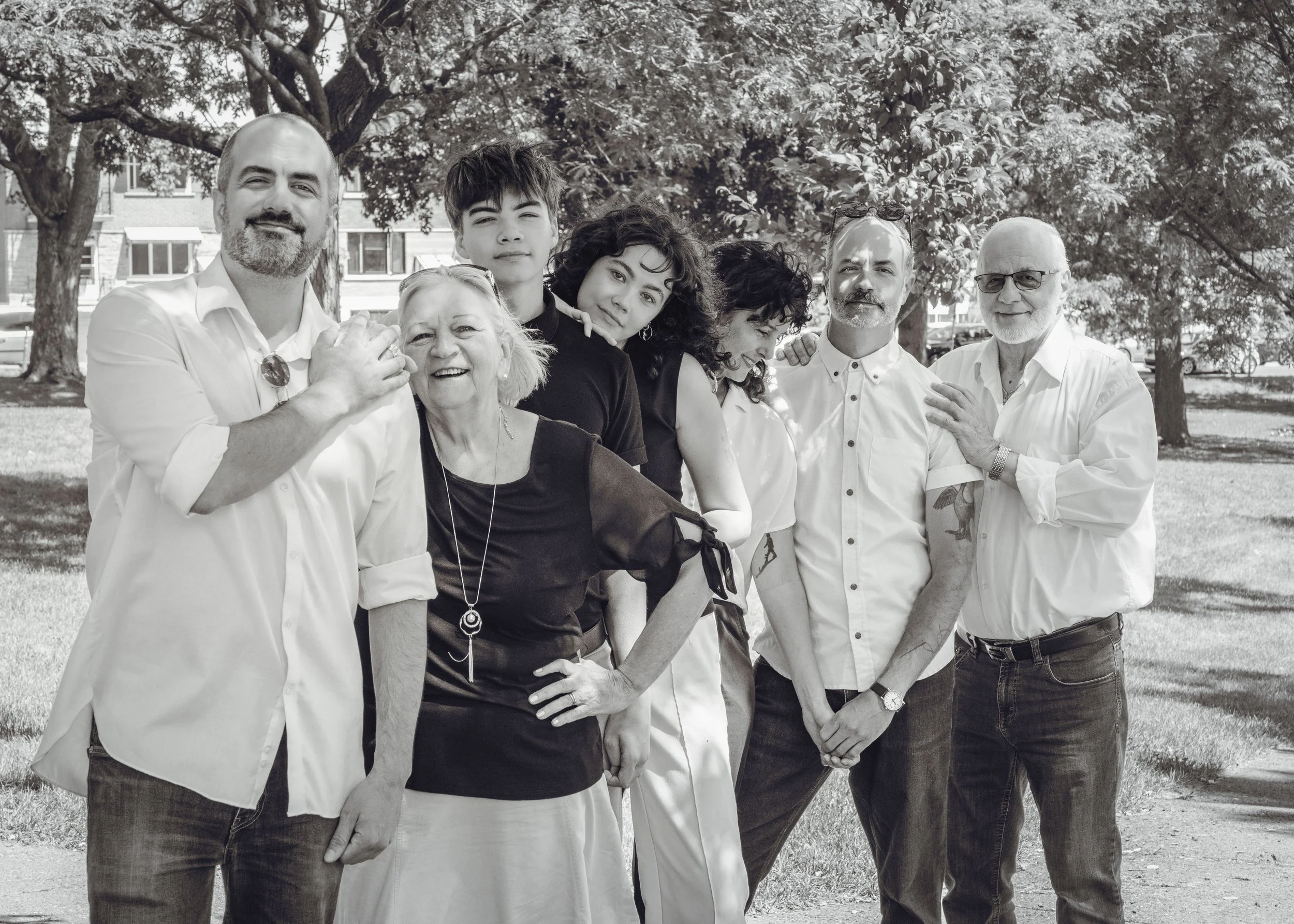 A group of eight diverse people standing outdoors under trees, smiling and posing together in a casual setting.